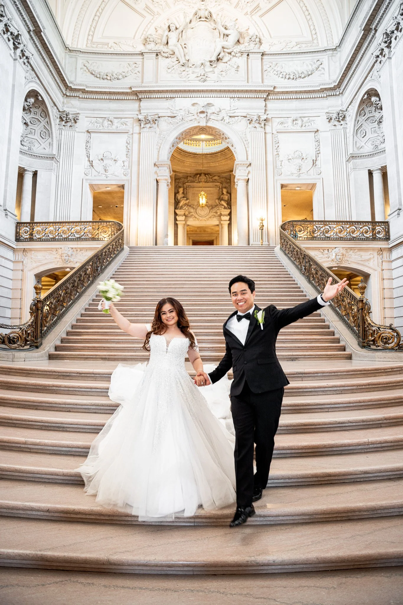 A newlywed couple celebrating at the top of a grand staircase inside San Francisco City Hall. 