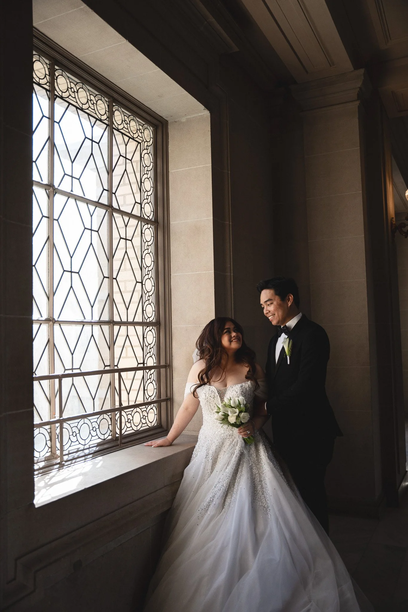A bride and groom standing by a large window with decorative ironwork, smiling at each other inside SF City Hall 1 Dr Carlton B Goodlett Pl, San Francisco, CA 94102.