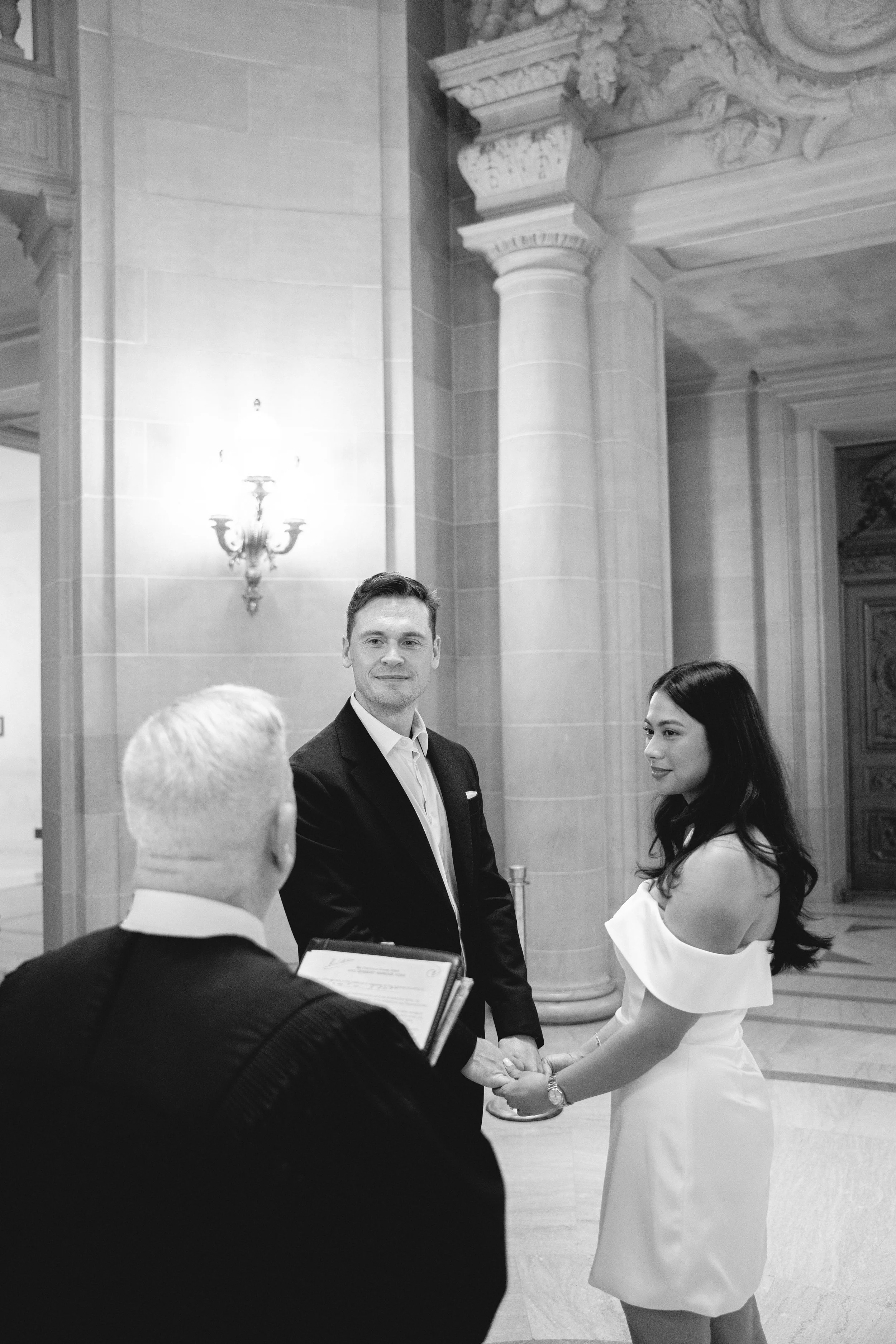 Couple exchanging vows before an officiant inside San Francisco City Hall's ceremony hall.