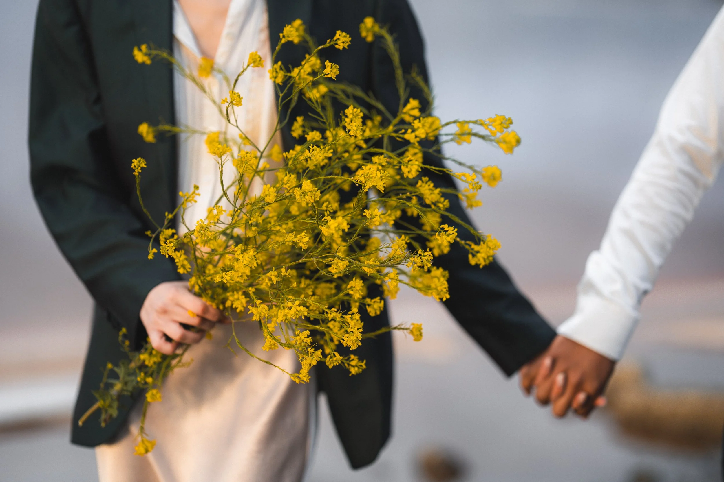 Two people holding hands, one person is holding a bouquet of yellow flowers. One person is wearing a dark suit and the other has a white sleeve visible, near pink lake in Marina Alviso.