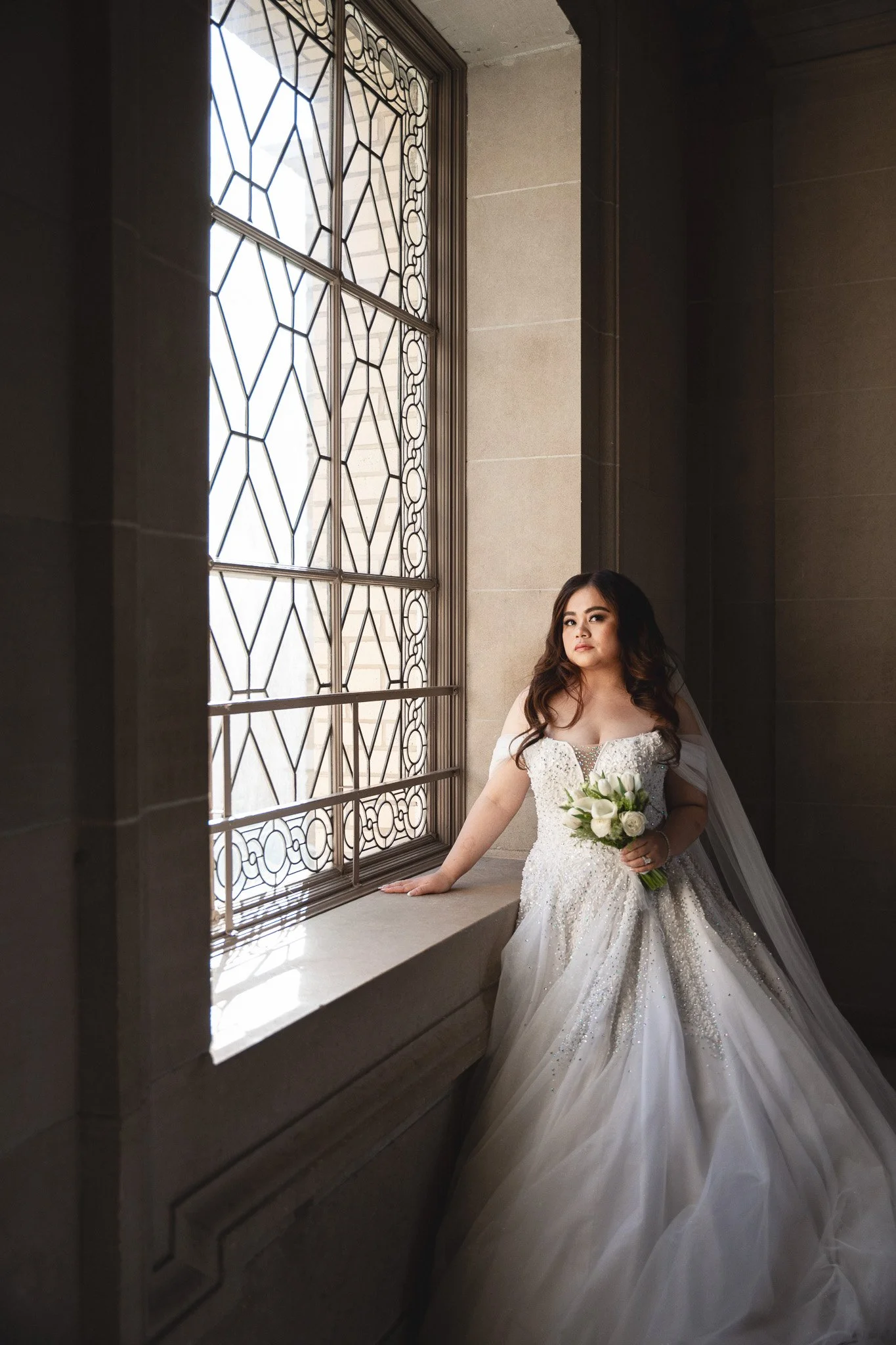 A bride in a white wedding gown holding a bouquet of white flowers, standing by a large decorative window with geometric patterns, in a stone-walled room inside San Francisco City Hall 1 Dr Carlton B Goodlett Pl, San Francisco, CA 94102.