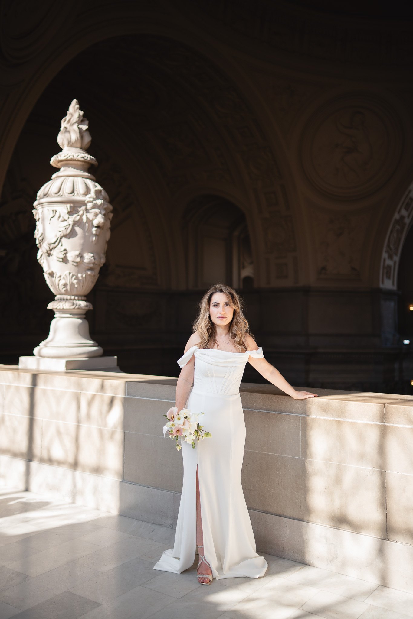 A woman in an off-shoulder white gown holding a bouquet standing on a stone ledge in an elegant interior with ornate architecture and large sculpture inside SF City Hall 1 Dr Carlton B Goodlett Pl, San Francisco, CA 94102.