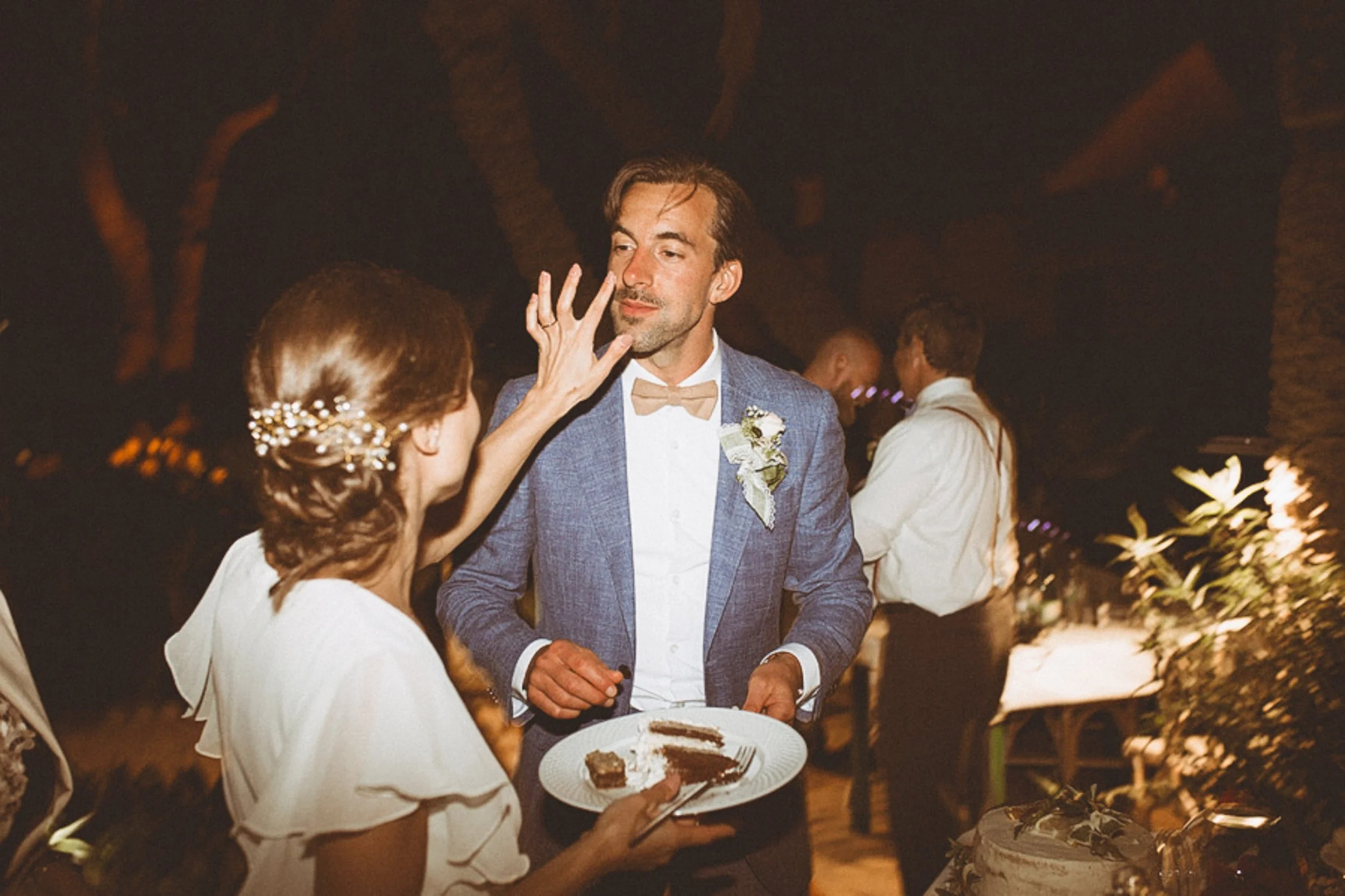 A man in a blue suit and beige bow tie at a wedding reception, reaching for a piece of chocolate cake held by a woman with brown hair in an elegant updo, secured with jeweled hairpins in a dimly lit venue decorated with flowers and tropical trees.