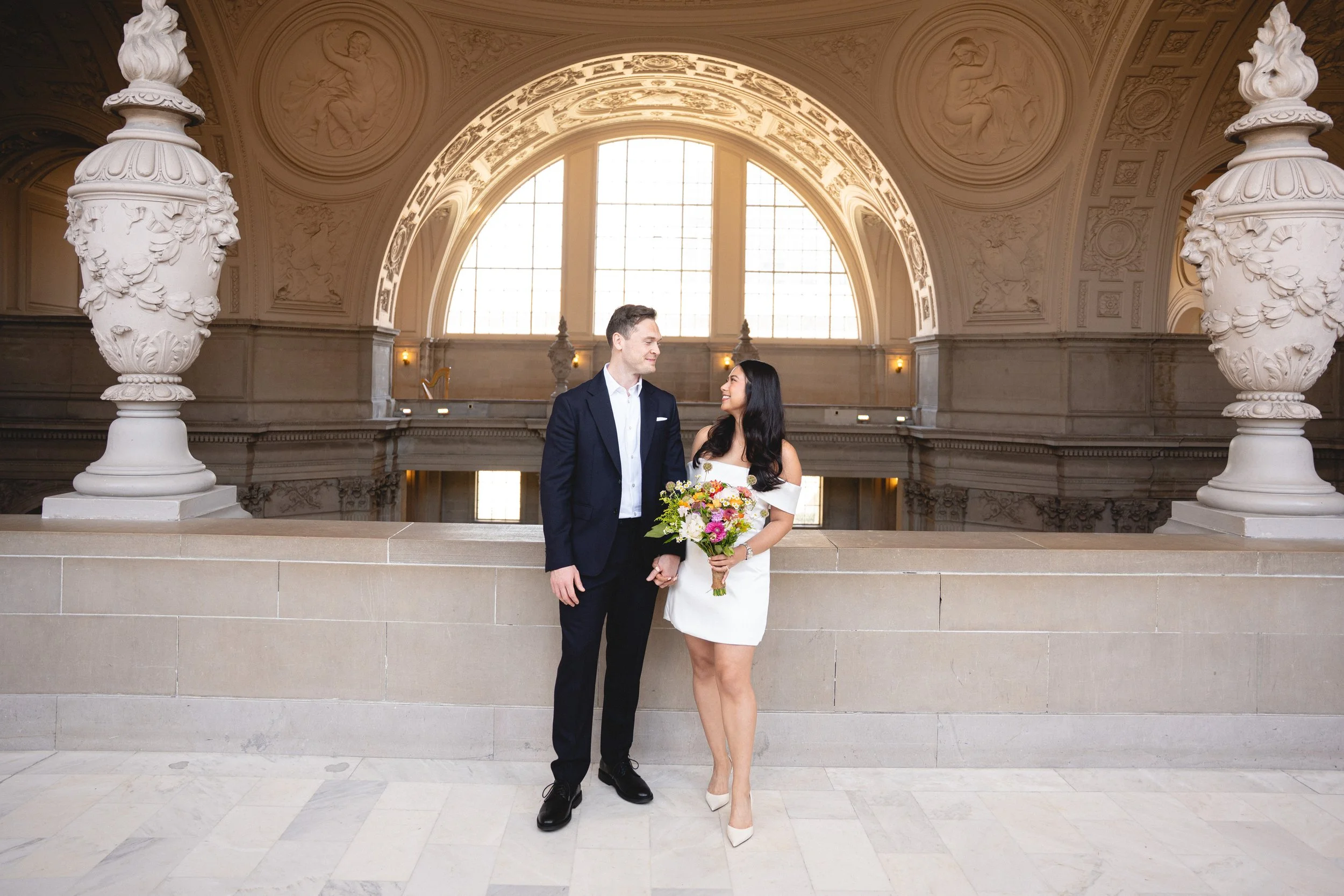 A couple dressed in wedding attire standing in front of a large arched window inside SF City Hall 1 Dr Carlton B Goodlett Pl, San Francisco, CA 94102. The woman is holding a bouquet of colorful flowers, and they are smiling at each other.
