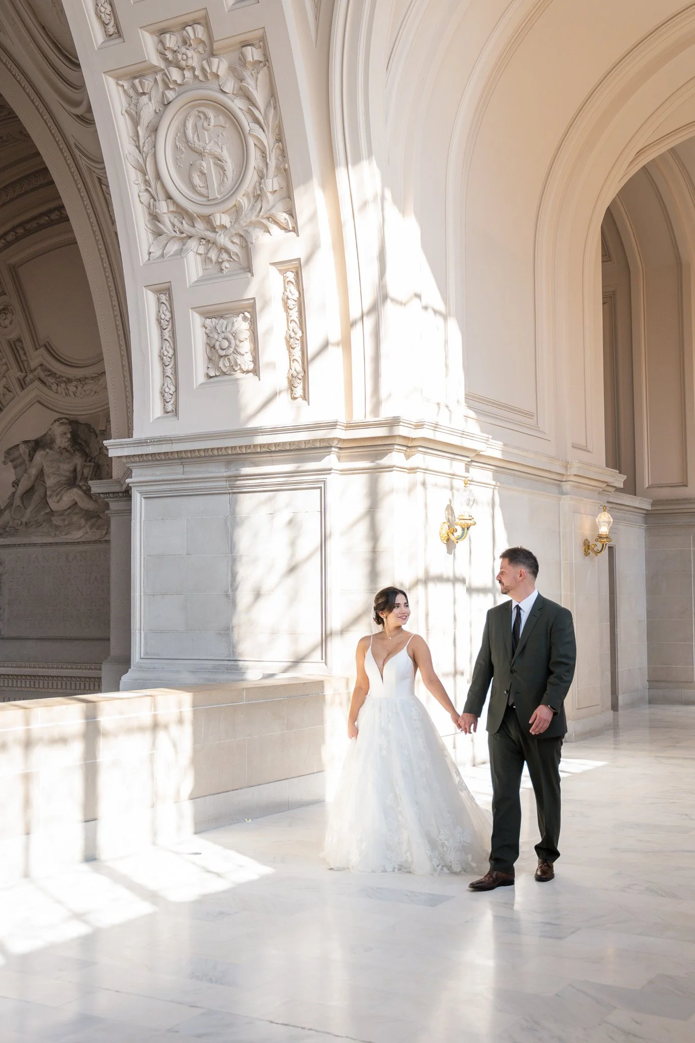 A bride and groom holding hands and walking inside San Francisco City Hall.. The bride wears a white wedding dress, and the groom wears a dark suit. Sunlight streams in, casting shadows on the marble floor.