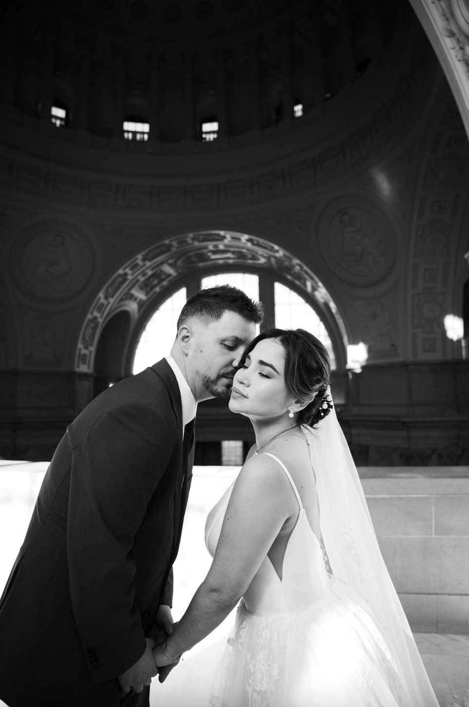 Black and white photo of a bride and groom holding hands and leaning in for a kiss inside San Francisco City Hall.
