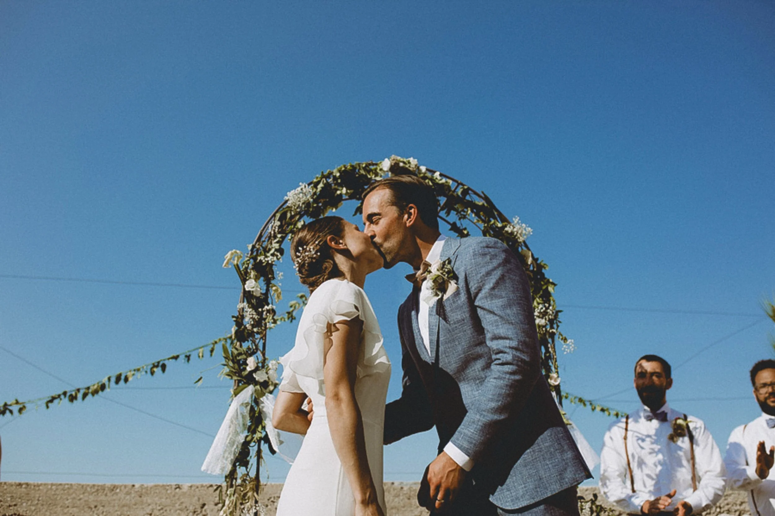 A couple is kissing under a floral arch at an outdoor wedding ceremony, with a clear blue sky in the background and wedding guests in the background.