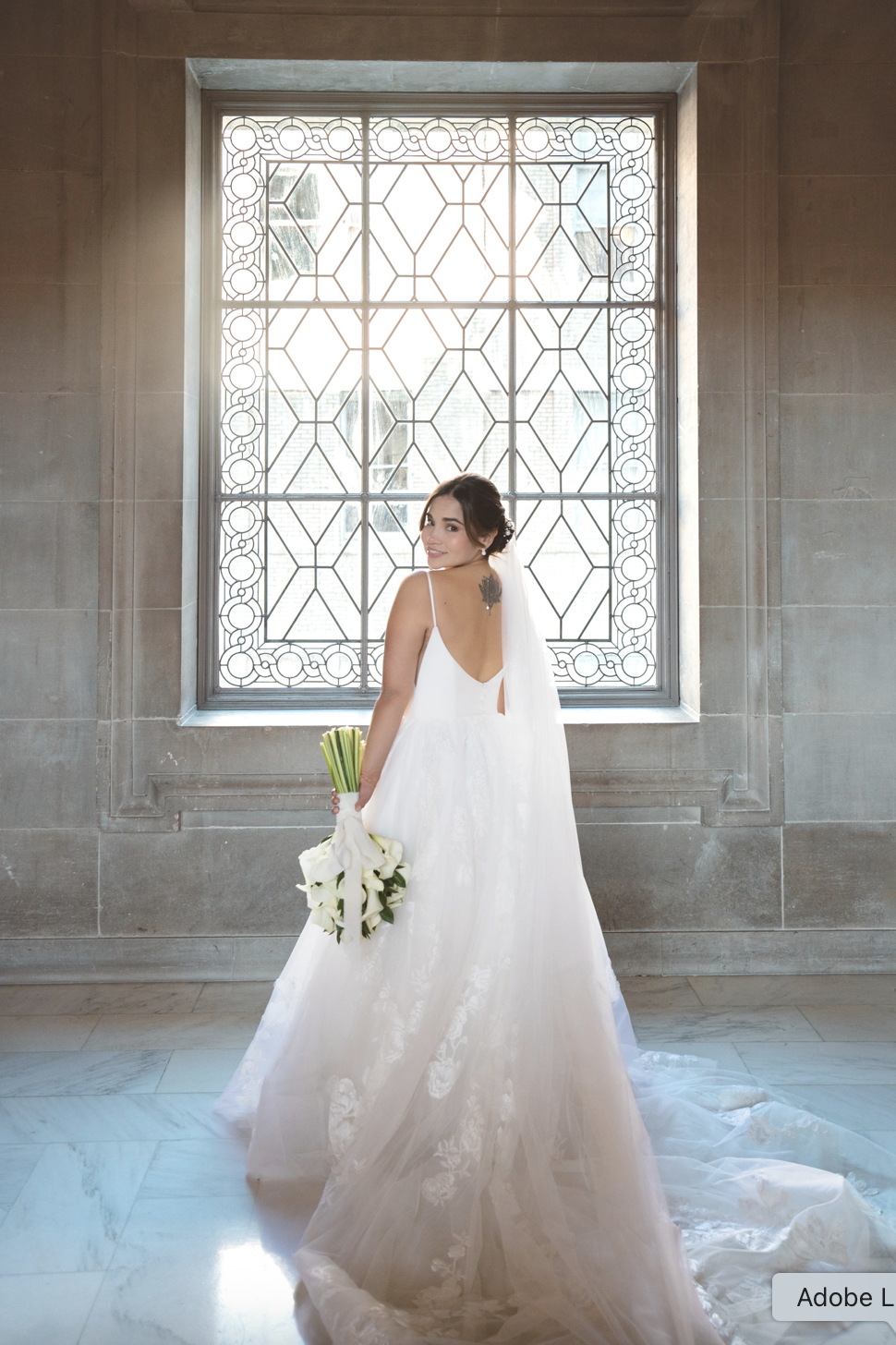 A bride in a white wedding gown holding a bouquet of white calla lilies, standing in front of a decorative window with geometric patterns, inside San Francisco City Hall.