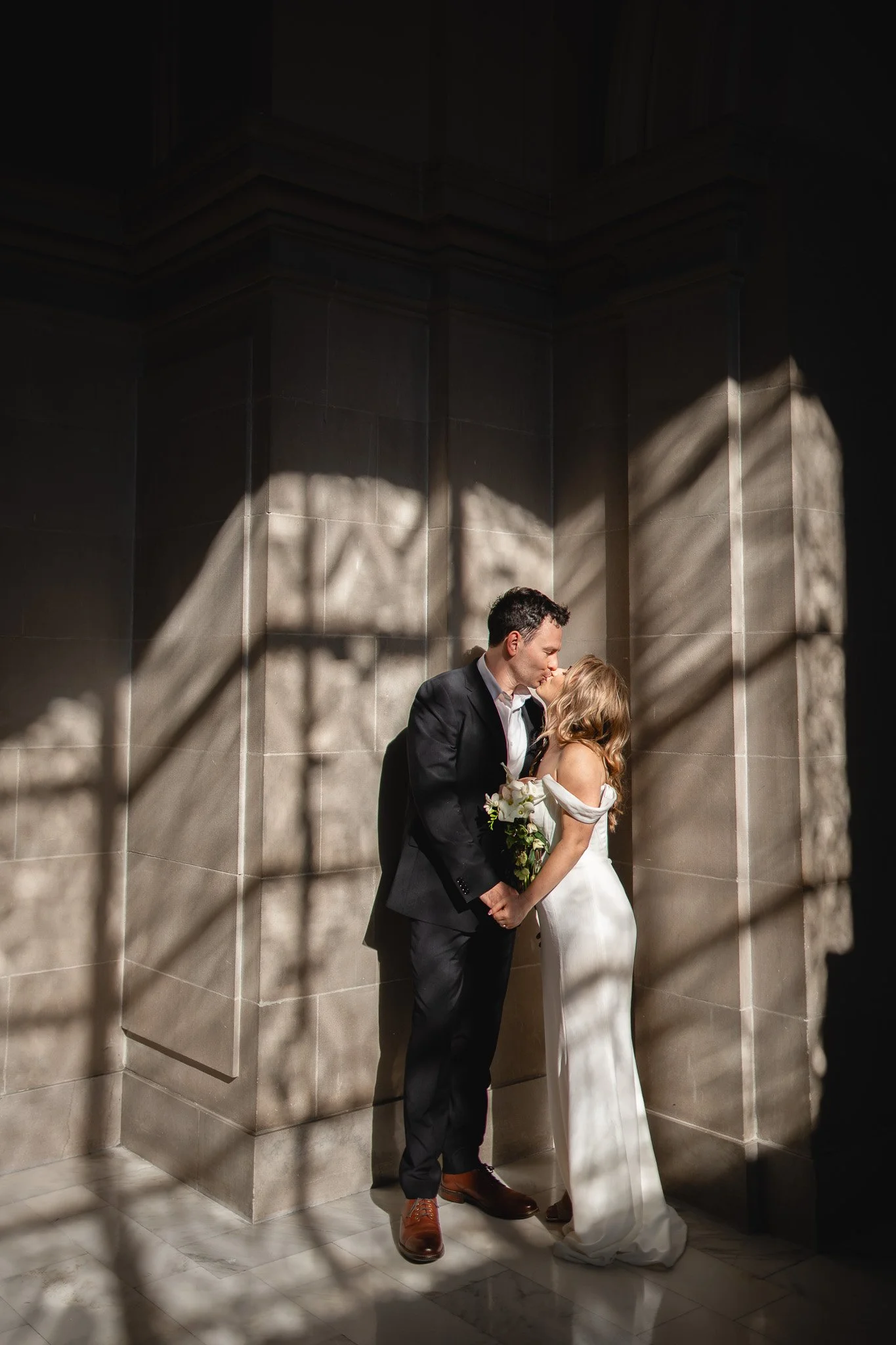 A couple in wedding attire sharing a kiss, holding hands, in a dimly lit room with shadows cast onto the wall inside San Francisco City Hall.
