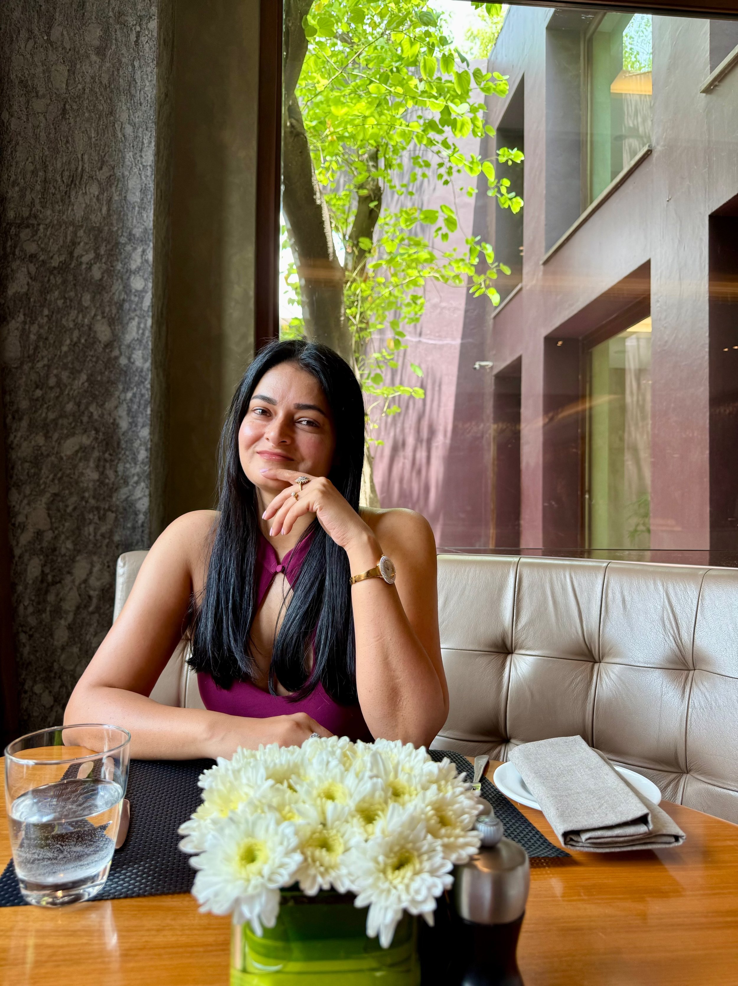 A woman with long dark hair, sitting at a table in a restaurant, smiling with her hand near her face, wearing a sleeveless maroon dress and jewelry. The table has a bouquet of white flowers, a glass of water, a napkin, and cutlery. The background shows a window with trees outside and modern building architecture.