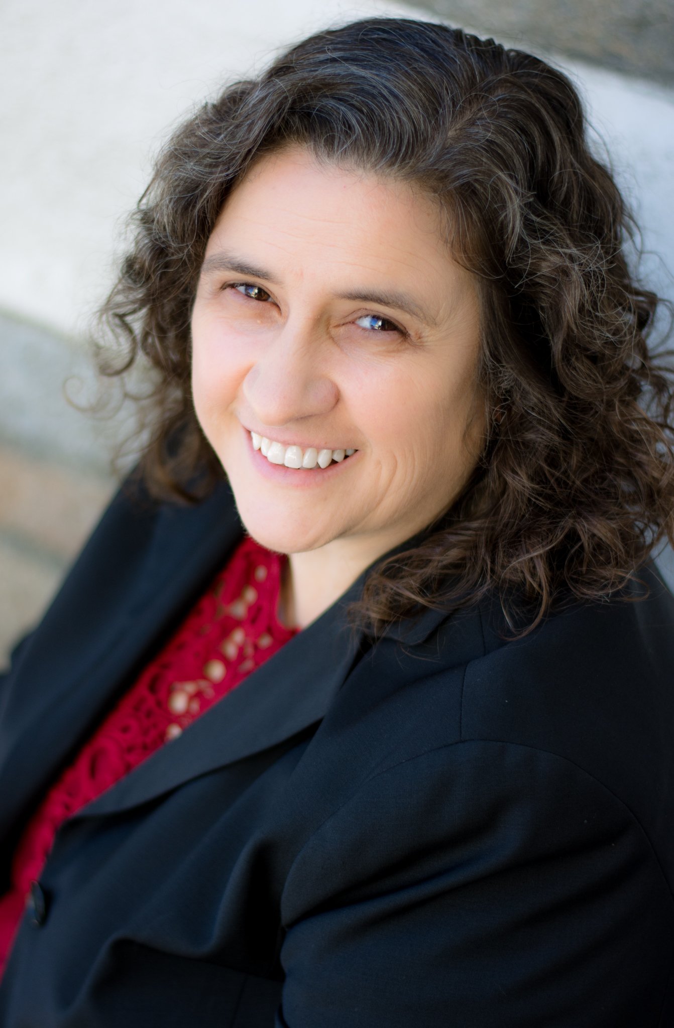 Close-up of a woman with curly dark hair smiling, wearing a black blazer and red top, outdoors.