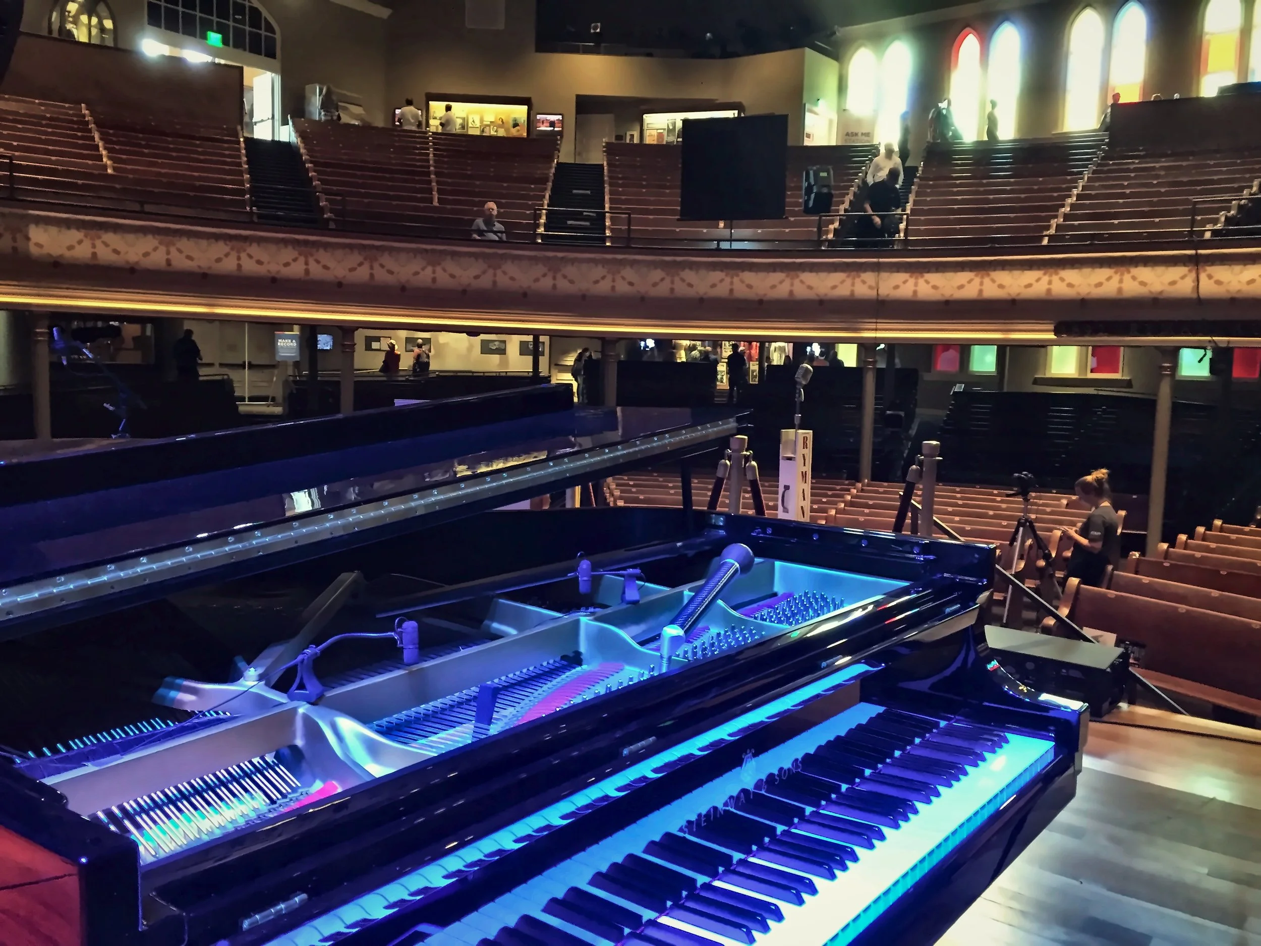 A grand piano with illuminated blue keys on a stage in a large auditorium, with multiple tiers of empty seating and a balcony, and a few people in the background preparing for a concert or event.