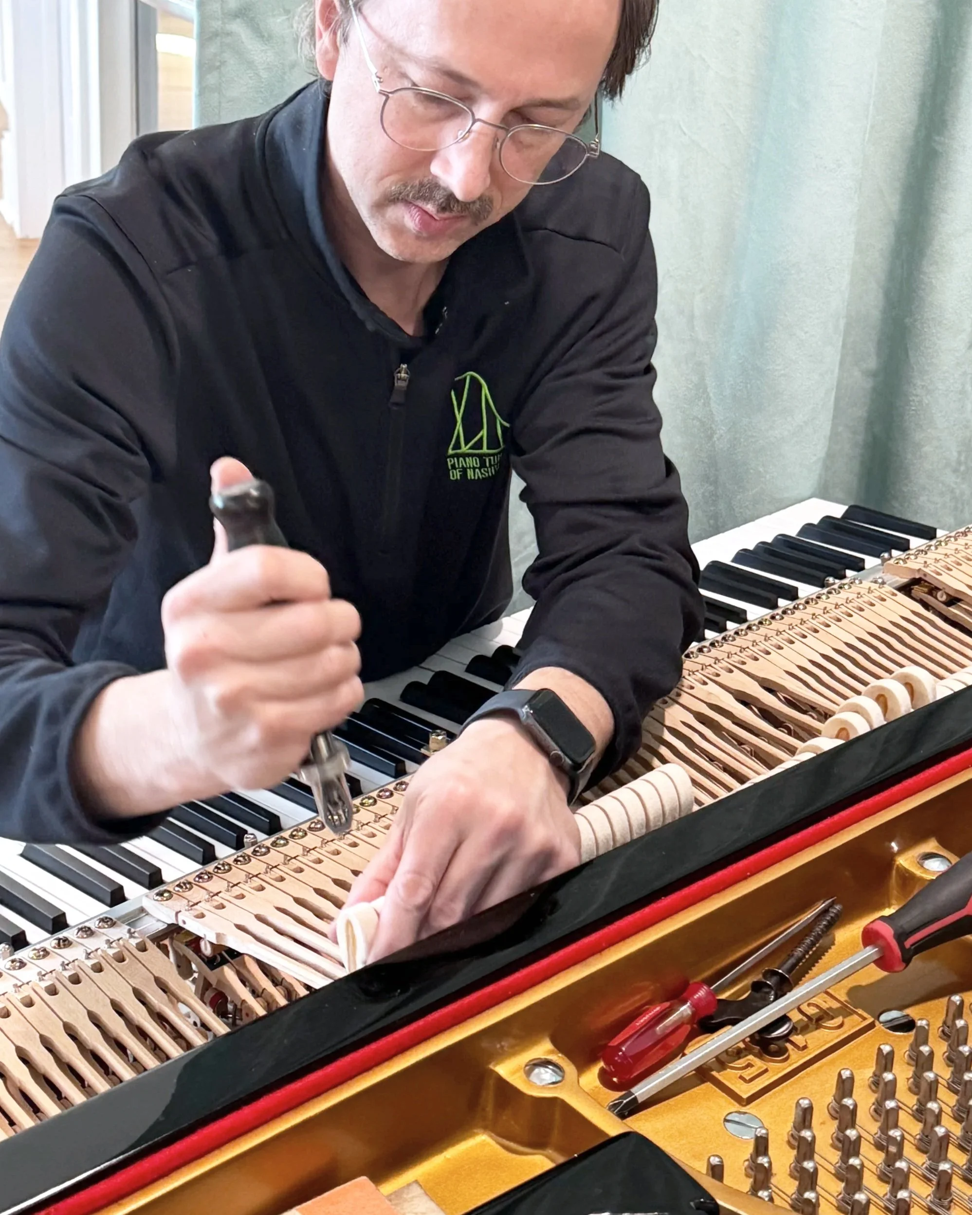 A man with glasses and a mustache working on a grand piano, focusing on voicing the piano hammers.