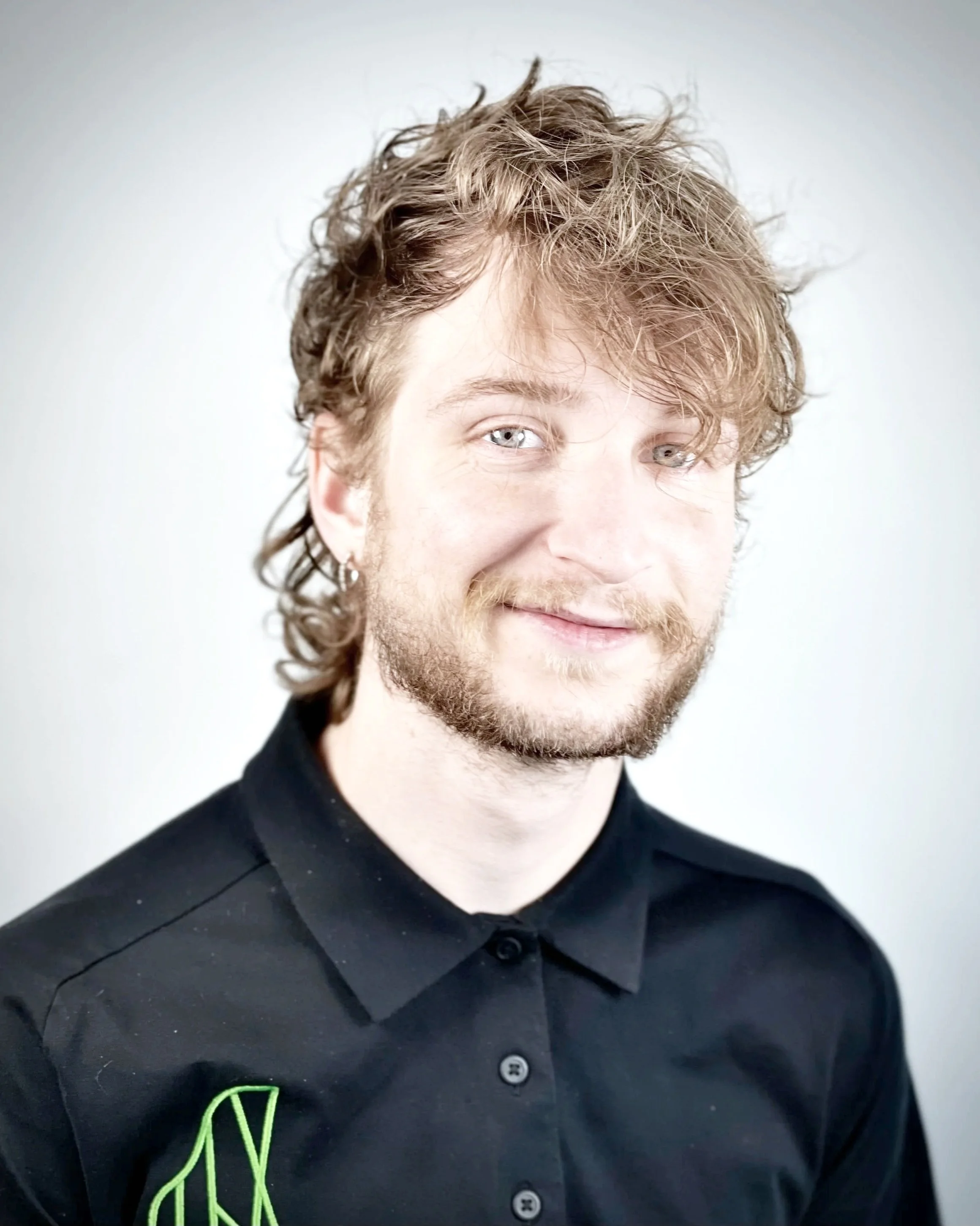 Close-up of a man with curly, light brown hair and a beard, wearing a black collared shirt with green embroidery on the chest.