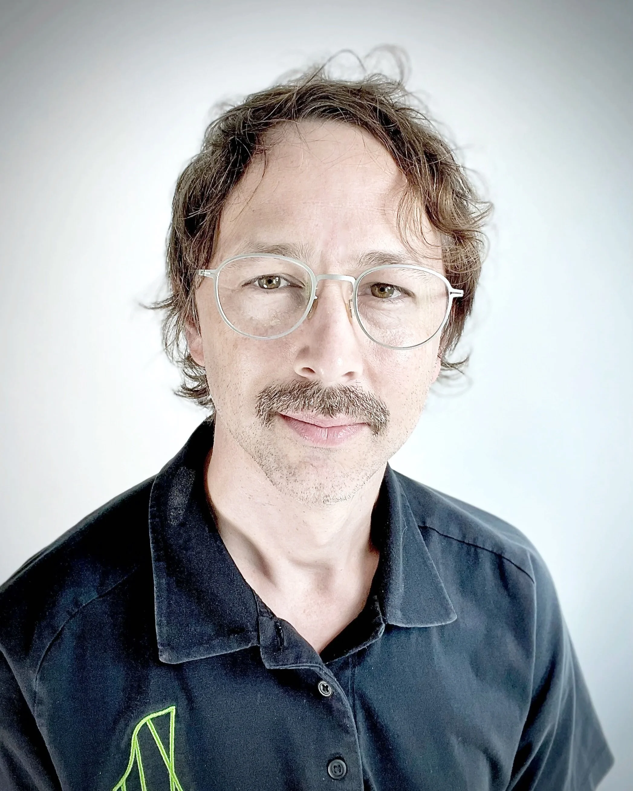 Close-up of a man with wavy brown hair, wearing round glasses and a black shirt with a lime green logo on the collar, against a plain white background.