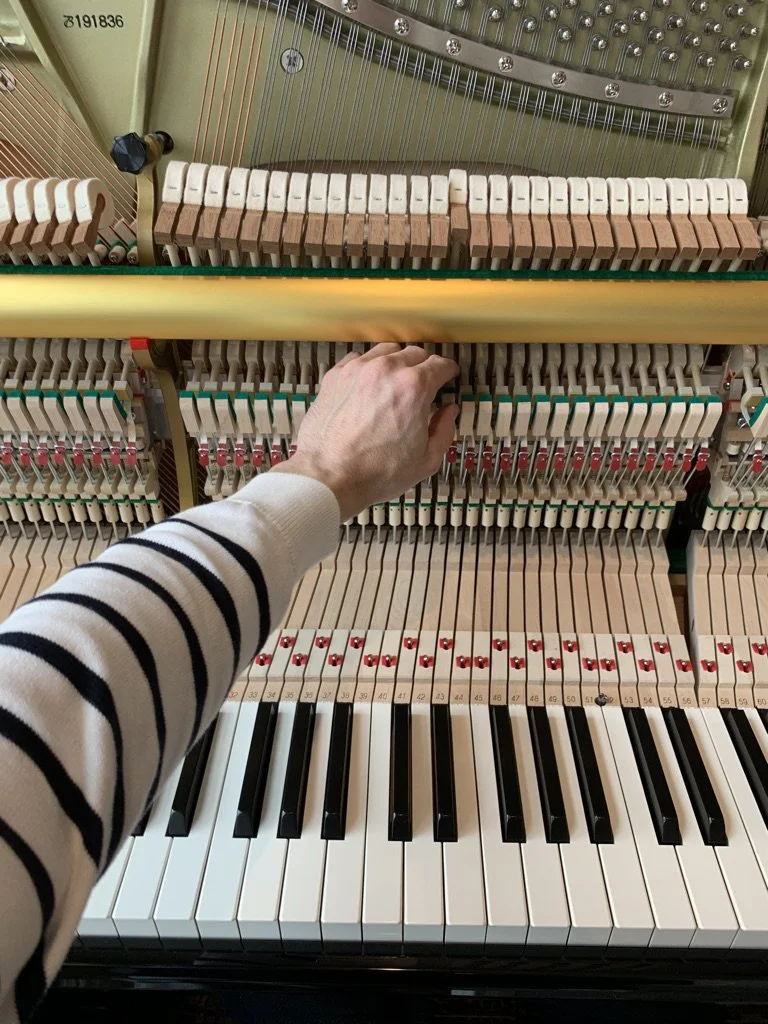 The interior of an upright piano showing the hammers, strings, and keys, with a person reaching inside to adjust or regulate the action.