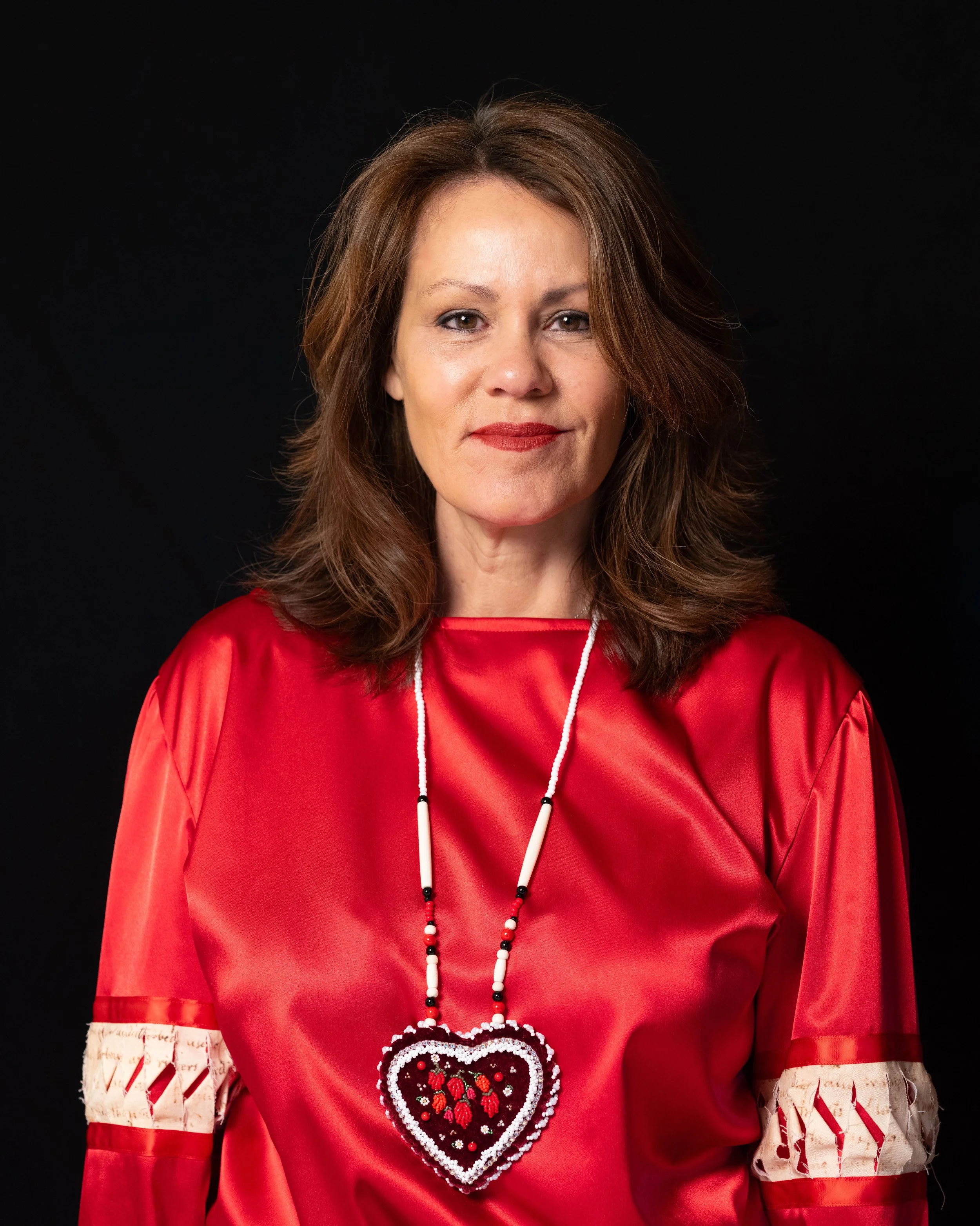 A woman wearing a red dress with white lace and red ribbon accents, a beaded necklace, and a large heart-shaped embroidered purse.