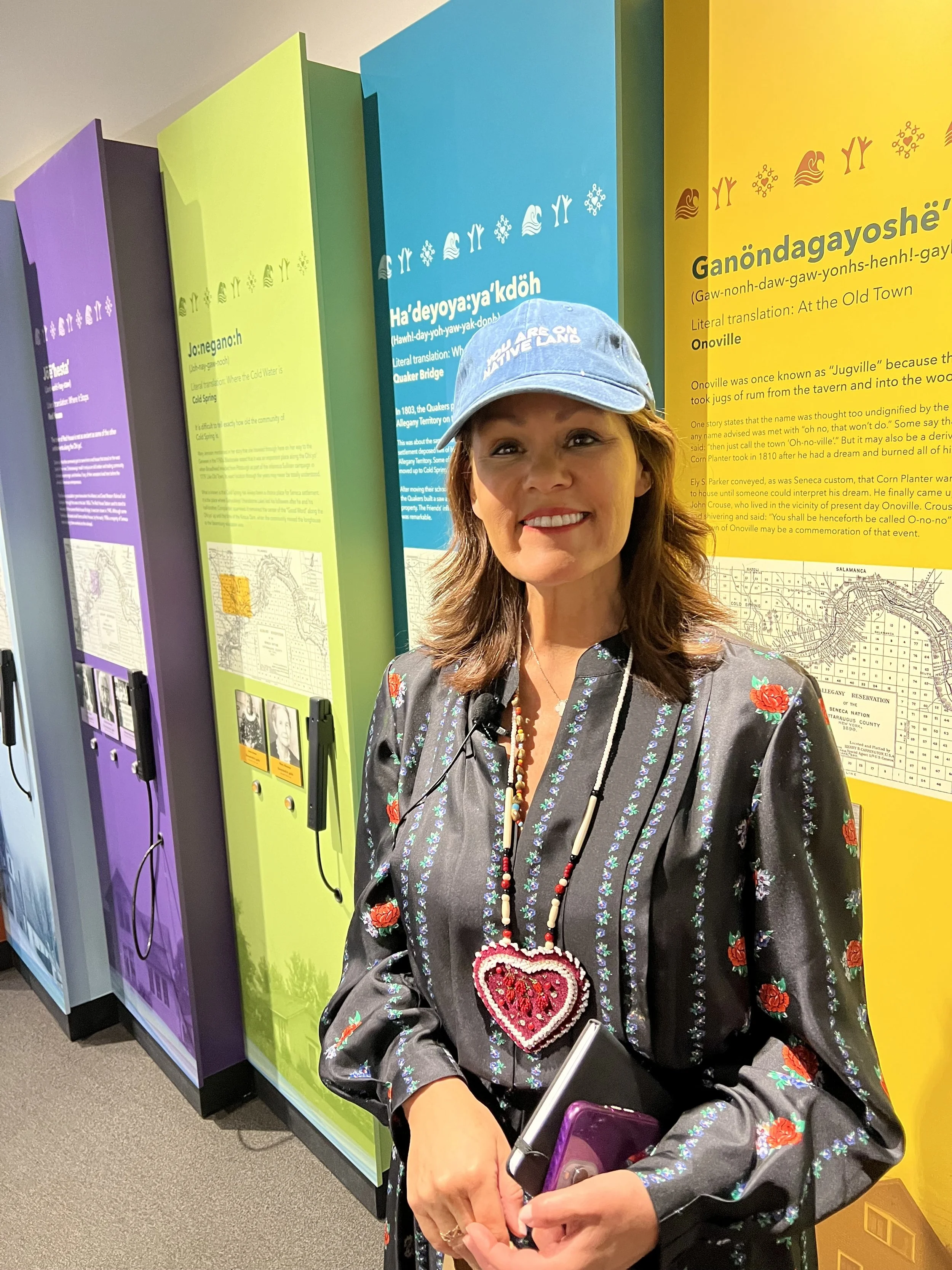 A woman with brown hair smiling while holding a smartphone, standing in front of colorful informational displays about Native American history and culture.