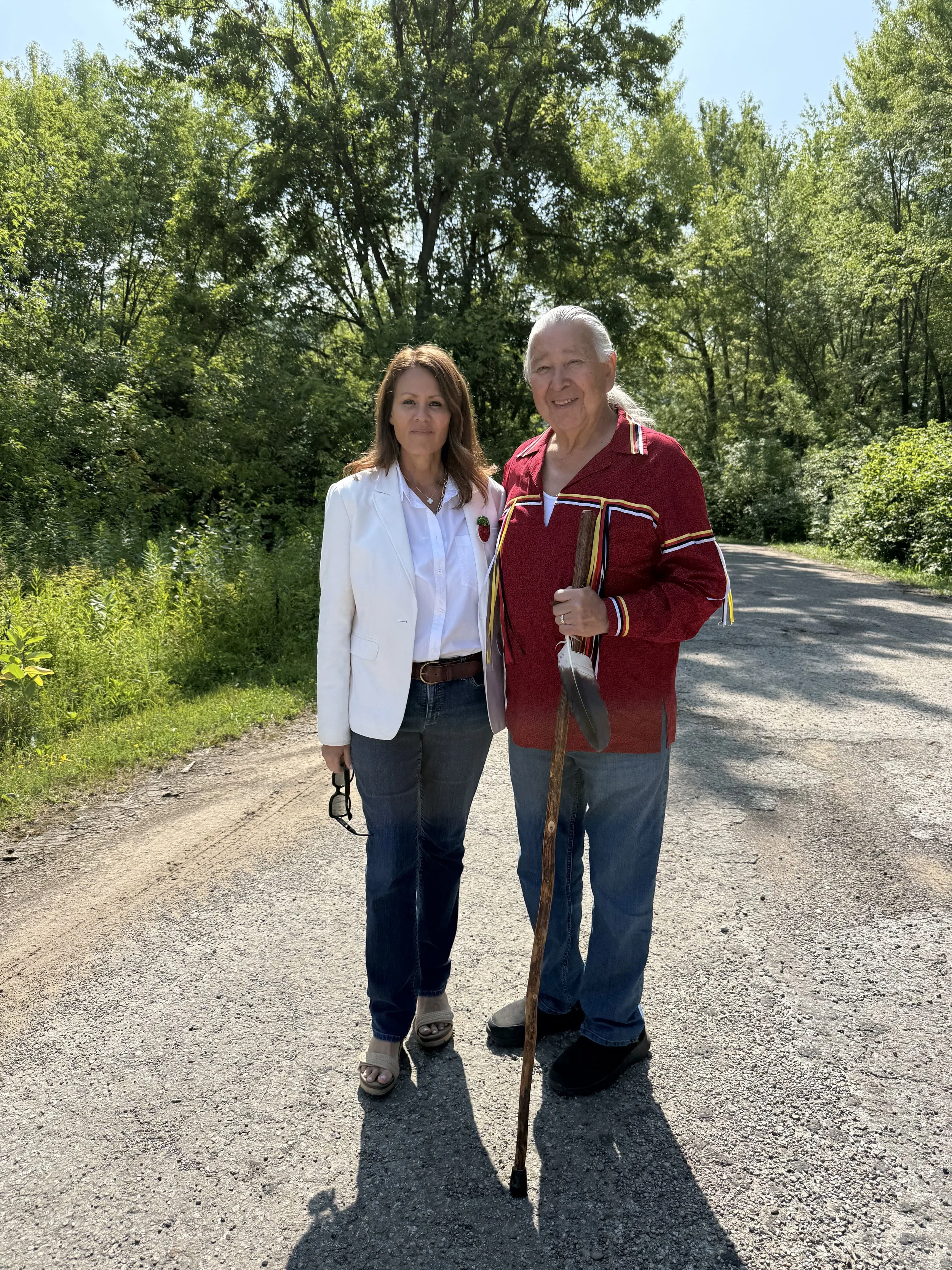 A woman and an elderly man standing outdoors on a gravel path surrounded by green trees. The woman is wearing a white blazer and dark jeans, and the man is dressed in a red jacket with white and yellow stripes, holding a walking stick, smiling at the camera on a sunny day.
