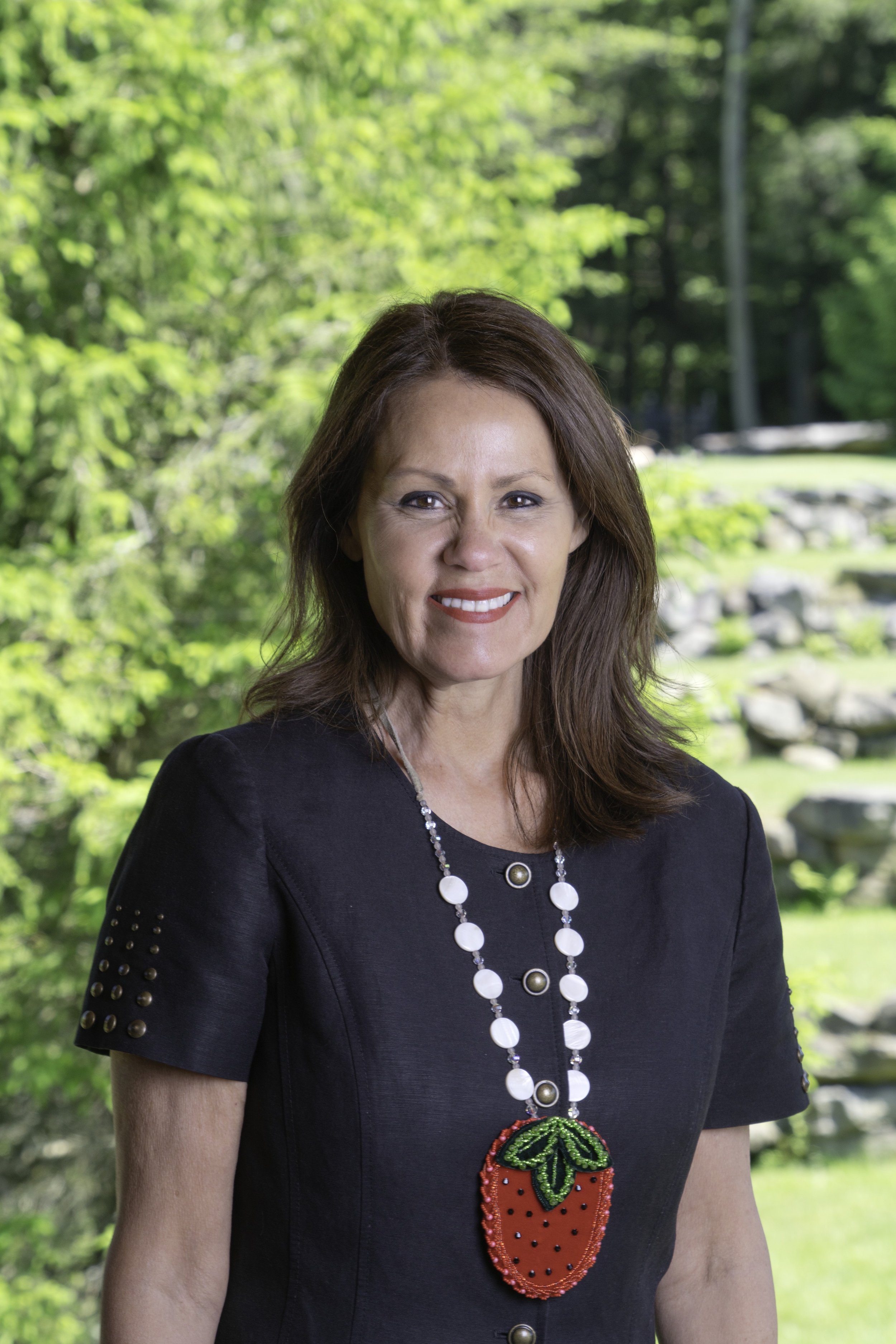 A woman with brown hair smiling, wearing a black dress with studded sleeves and a white beaded necklace with a strawberry-shaped pendant, outdoors with green trees and rocks in the background.