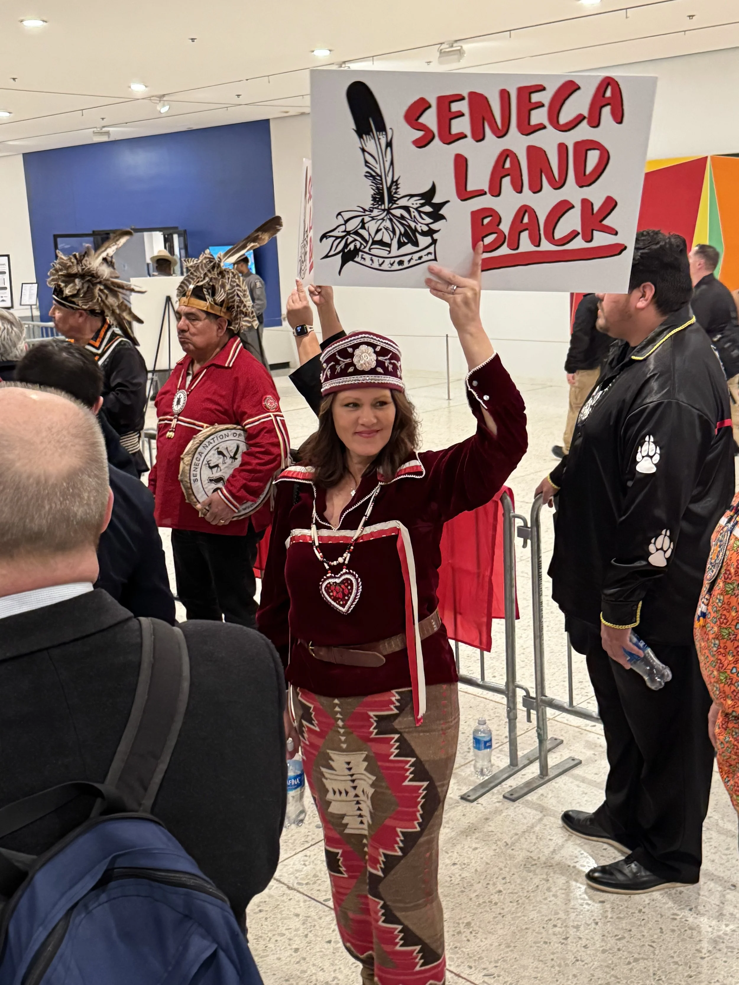 A woman holding a sign that reads "Seneca Land Back" at a protest or rally, surrounded by other people including individuals in traditional indigenous clothing and headwear, in what looks like an indoor public space.