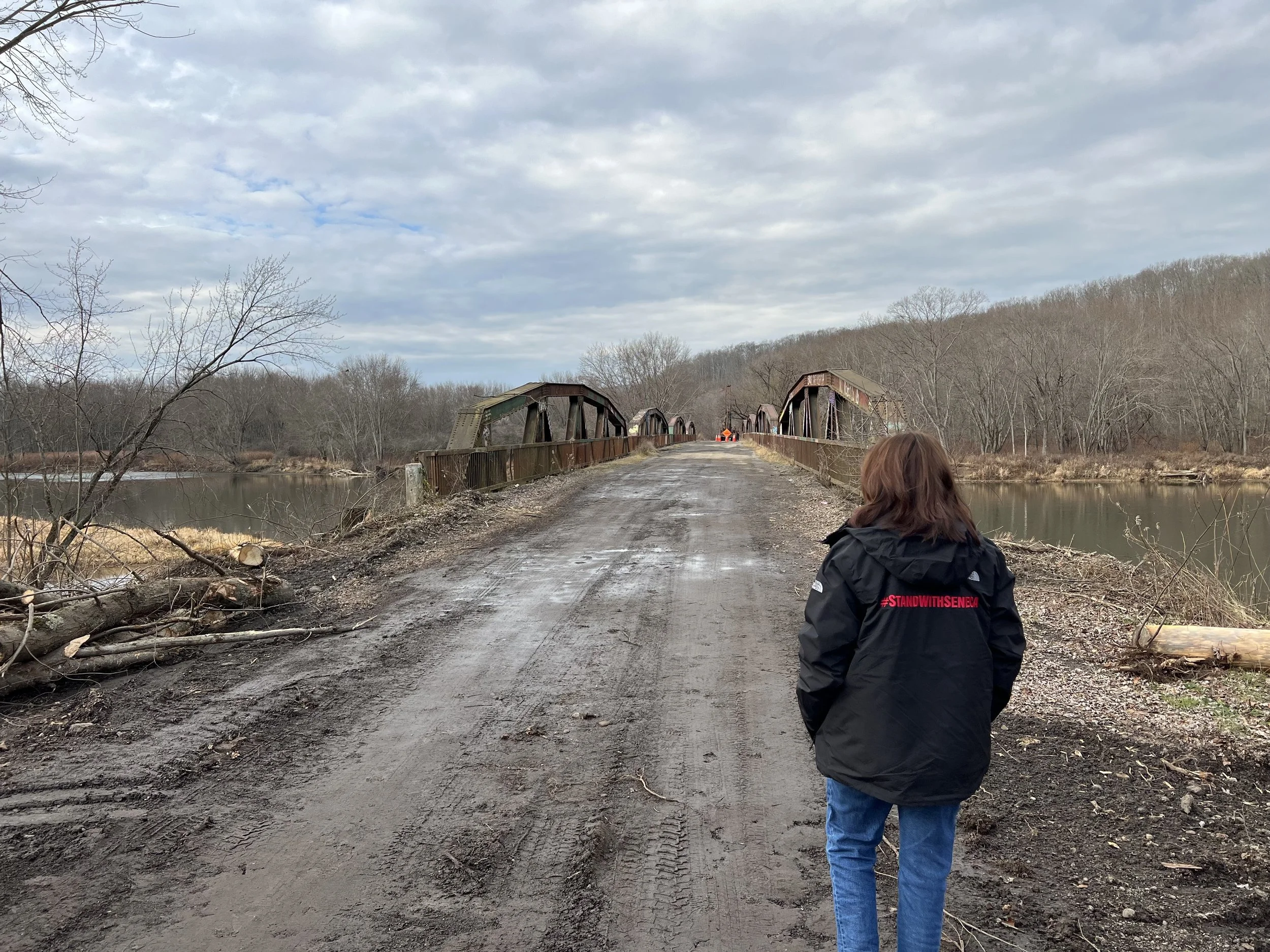 A woman wearing a black jacket with red text '#STANDWITHSENEA' on the back stands on a muddy dirt road near a river, facing an old, rusty metal bridge over the river. Trees without leaves line both sides of the river, and the sky is partly cloudy.