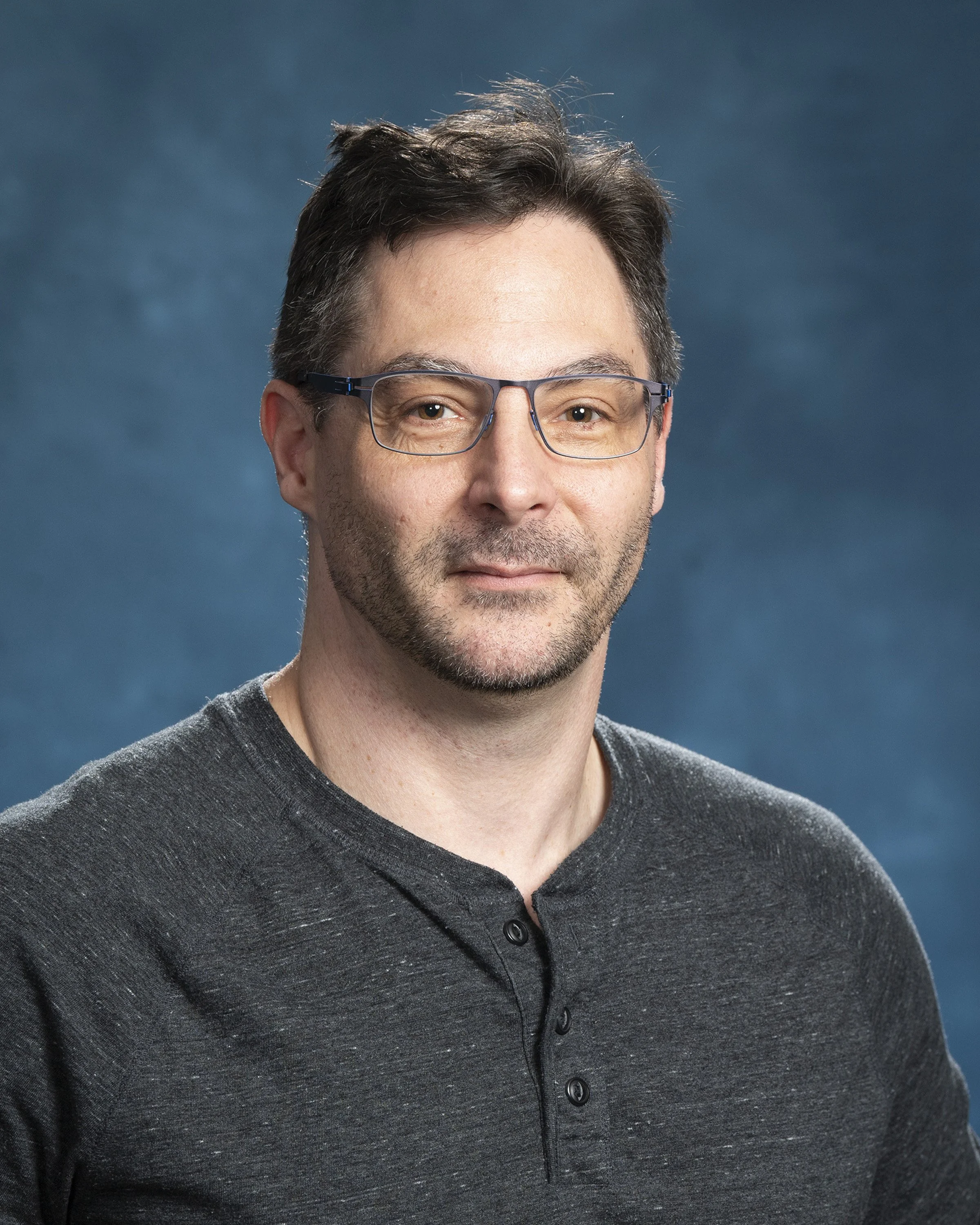Close-up of a man with glasses and dark hair, wearing a dark gray button-up shirt, against a blue gradient background.