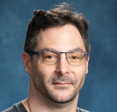 Close-up of a man with glasses and dark hair, smiling slightly, against a blue background.