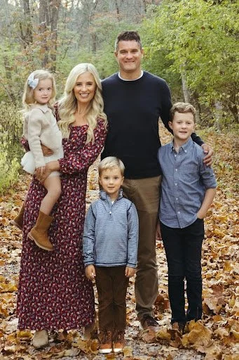 Family of five standing outdoors in a wooded area with fallen leaves, including a woman, man, and three children, smiling at the camera.