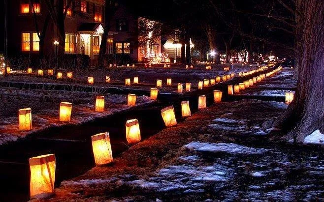 Night scene with rows of glowing lanterns along a snow-covered pathway, with trees and buildings illuminated in the background.