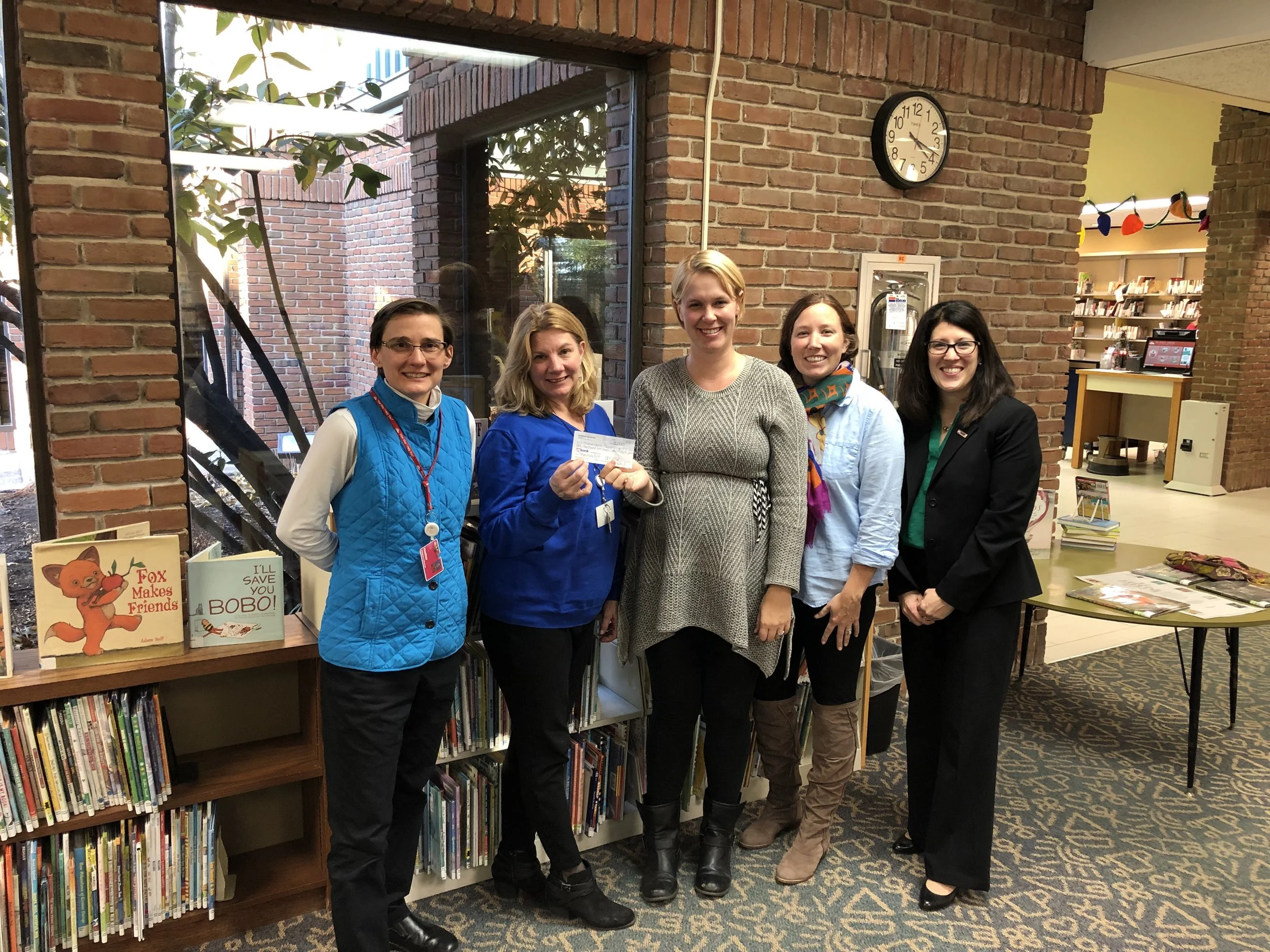 Five women standing together in a library, smiling at the camera. There are bookshelves behind them, and a brick wall with a clock showing 2:12. Two of the women are holding check or paper, and one woman is wearing a badge around her neck.