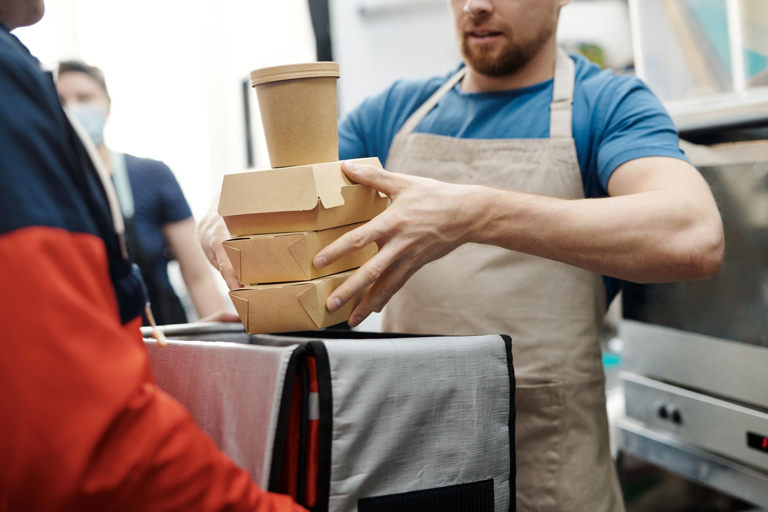Person wearing a blue shirt and apron handing over stacked takeout food containers and a coffee cup to another person at a food counter.