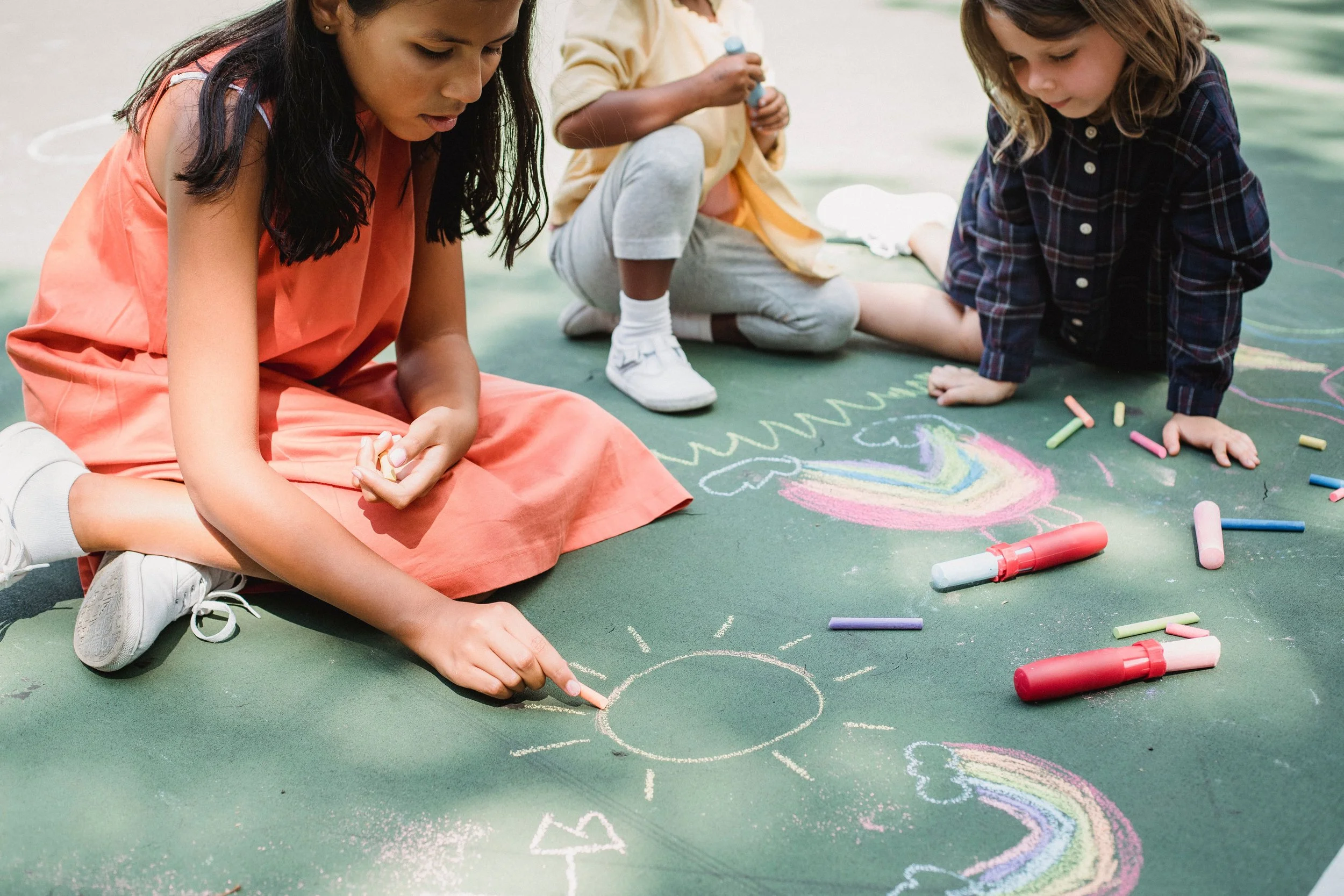 Children drawing with chalk on a green outdoor surface, creating colorful rainbow and heart shapes.