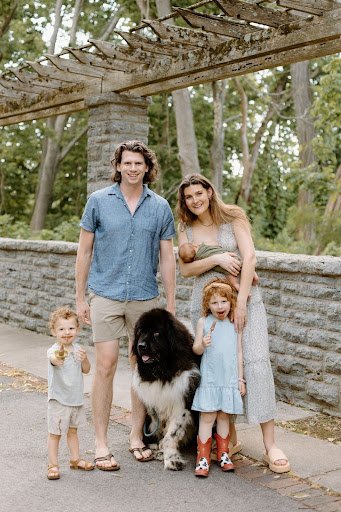 A family of four with a large black and white dog standing outdoors on a sidewalk. The father is wearing a blue short-sleeve shirt and shorts, the mother is in a light-colored dress holding a young girl, and there are two small children, a boy and a girl, standing nearby. They are in a park with trees and a stone wall in the background.