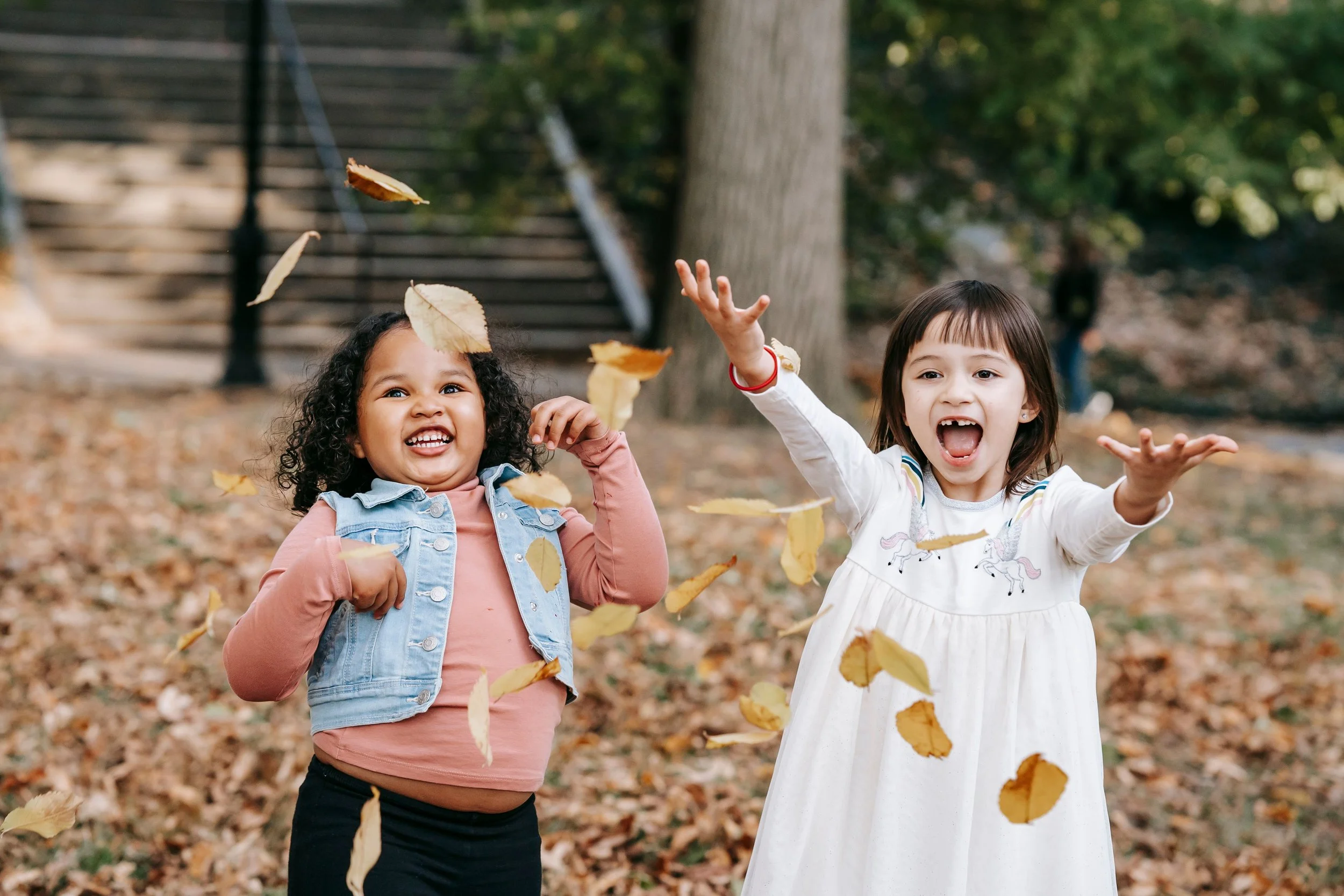 Two young girls playing with falling autumn leaves in a park, with trees and stairs in the background.