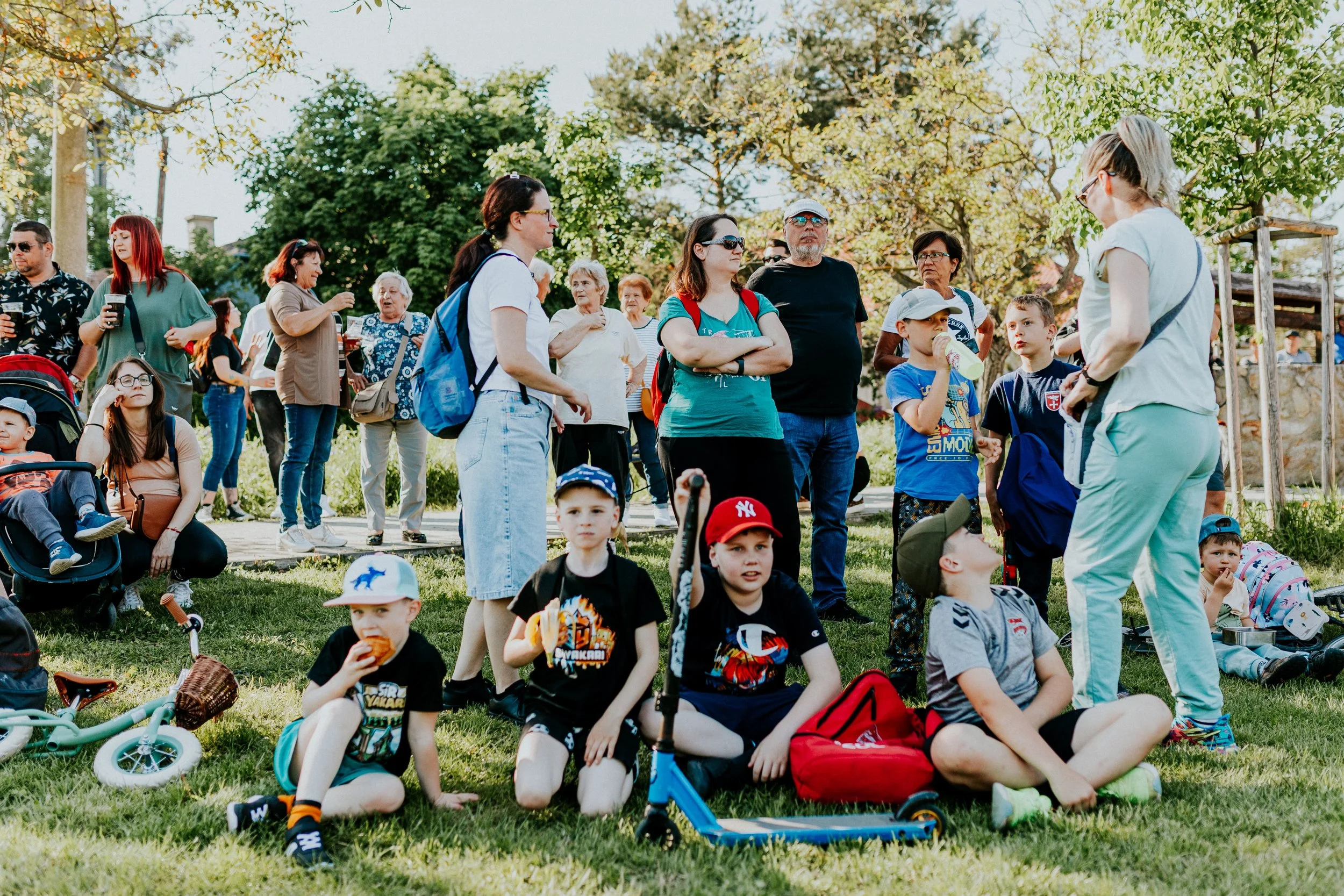 A group of children and adults gathered in a park on a sunny day, some sitting on the grass and others standing, with trees in the background. The children are seated on the grass with toys and backpacks, while adults are standing or sitting nearby, engaged in conversation.
