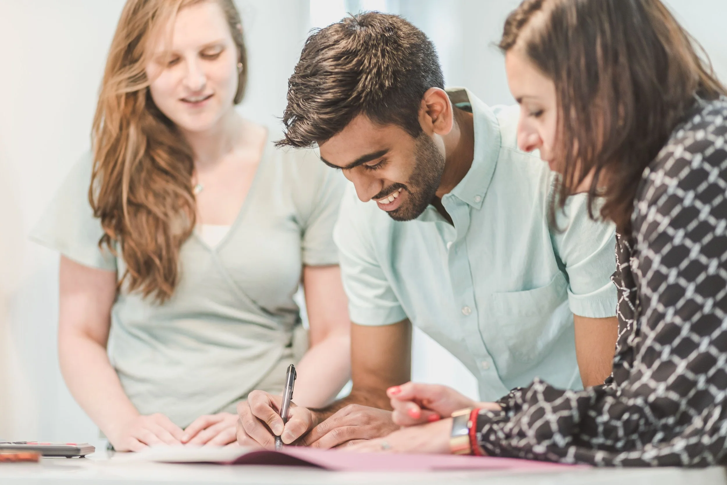 Three people are gathered around a table, looking at papers and writing. They appear to be collaborating or working together on a project.