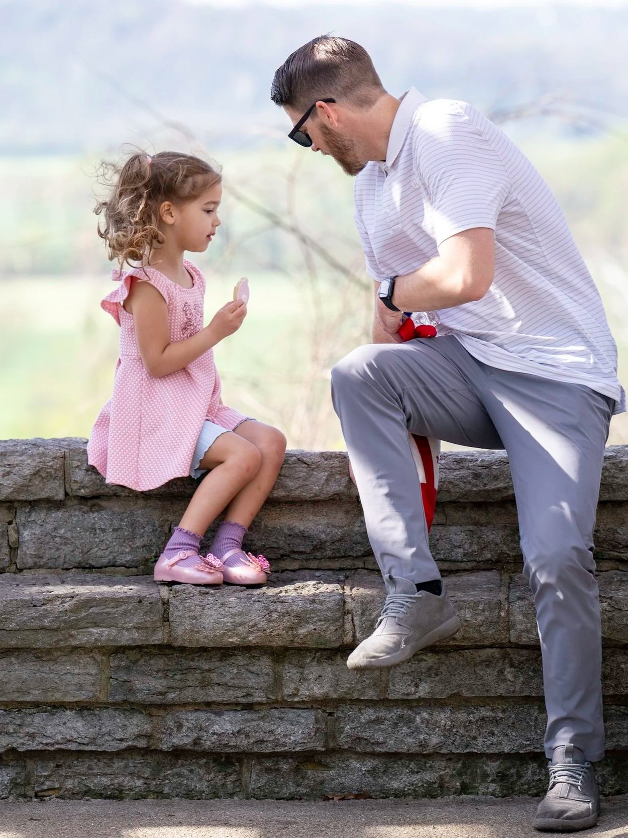 A man in a white striped polo shirt and gray pants kneeling on one knee, talking to a young girl sitting on a stone ledge. The girl is holding a pink lollipop, wearing a pink dress, purple socks, and pink shoes. The background is outdoors with a soft-focus green landscape.