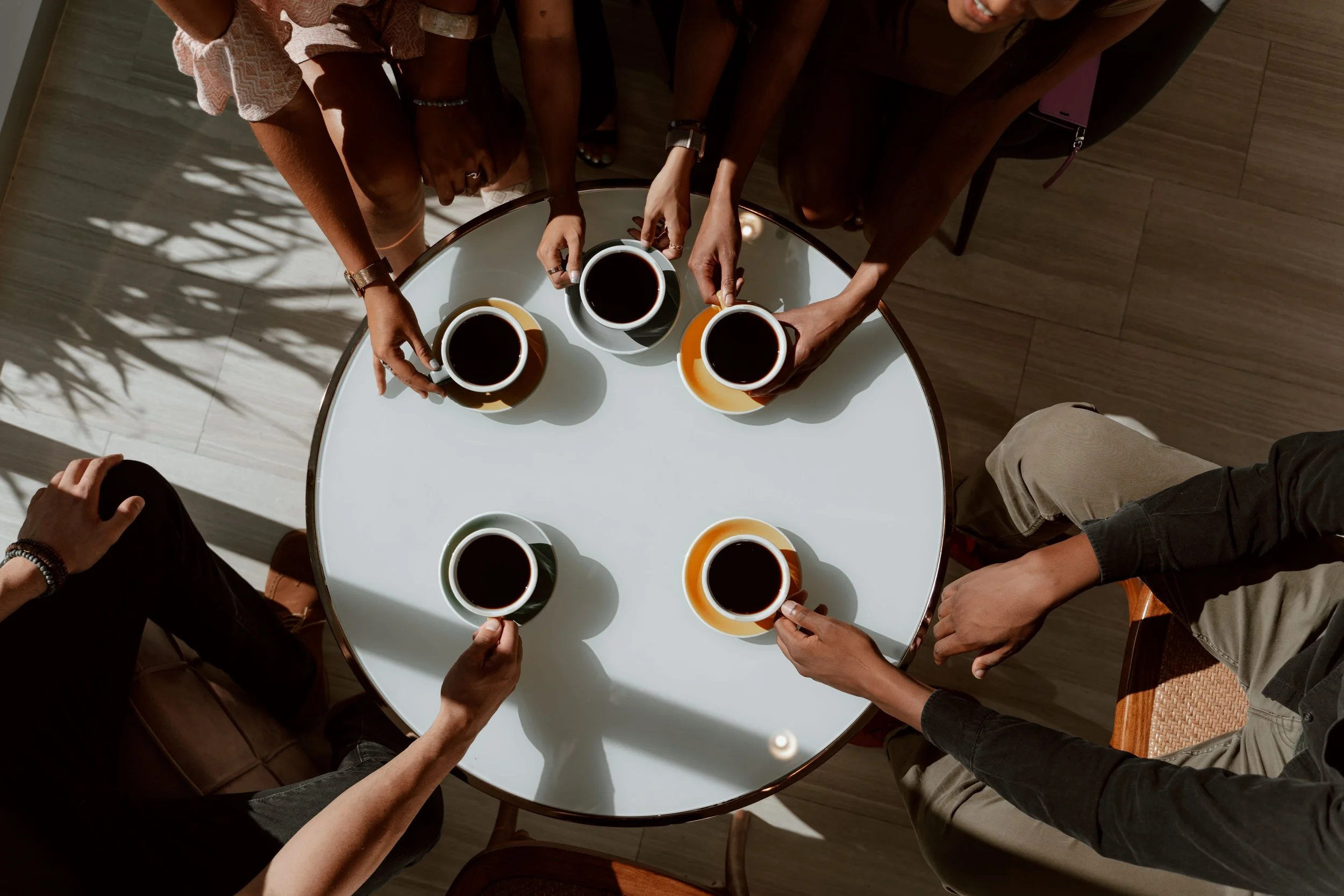 Six people sitting around a white table holding coffee cups from above in natural lighting.