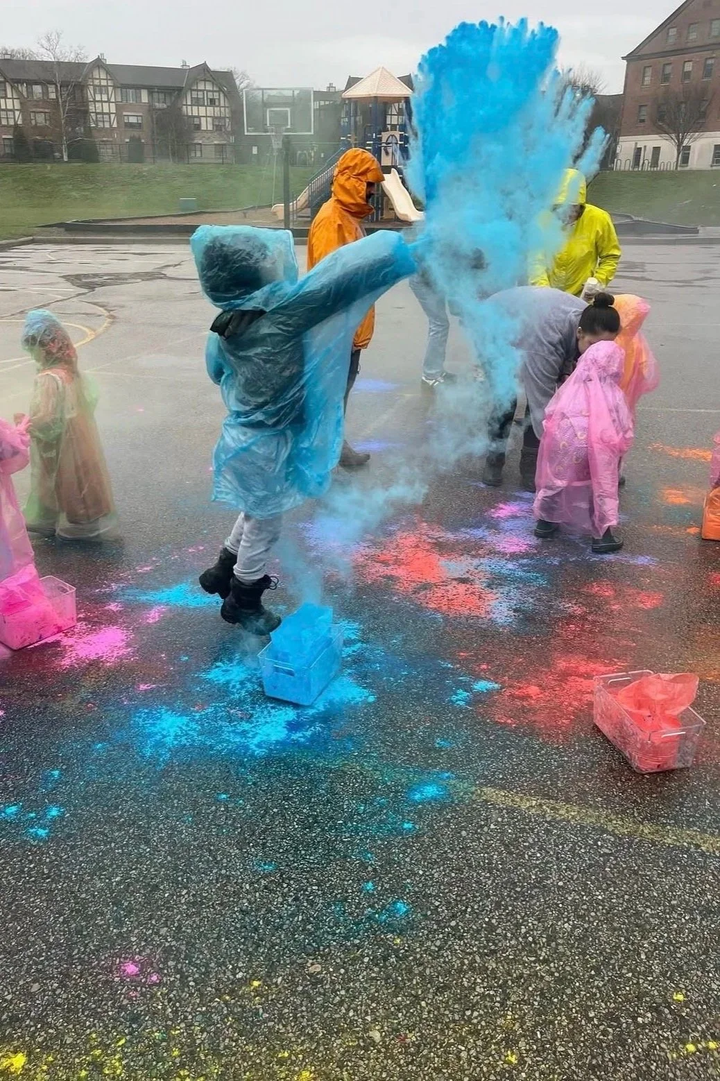 Children and adults celebrating the Holi festival outdoors on a rainy day, throwing colored powder and water. Participants wear rain ponchos in various colors, with colorful powders and splashes on the ground.