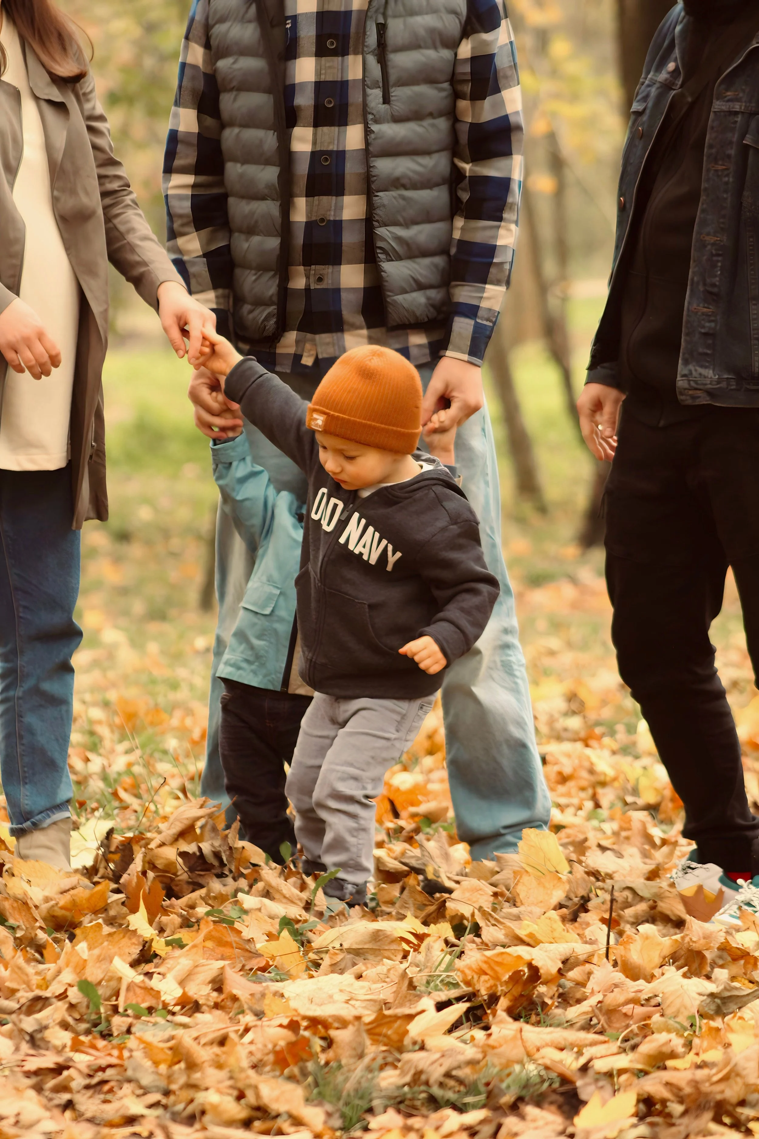 A young child wearing a brown beanie and a black hoodie with white lettering, holding hands with an adult while walking through a park covered in fallen autumn leaves, with other adults surrounding them.