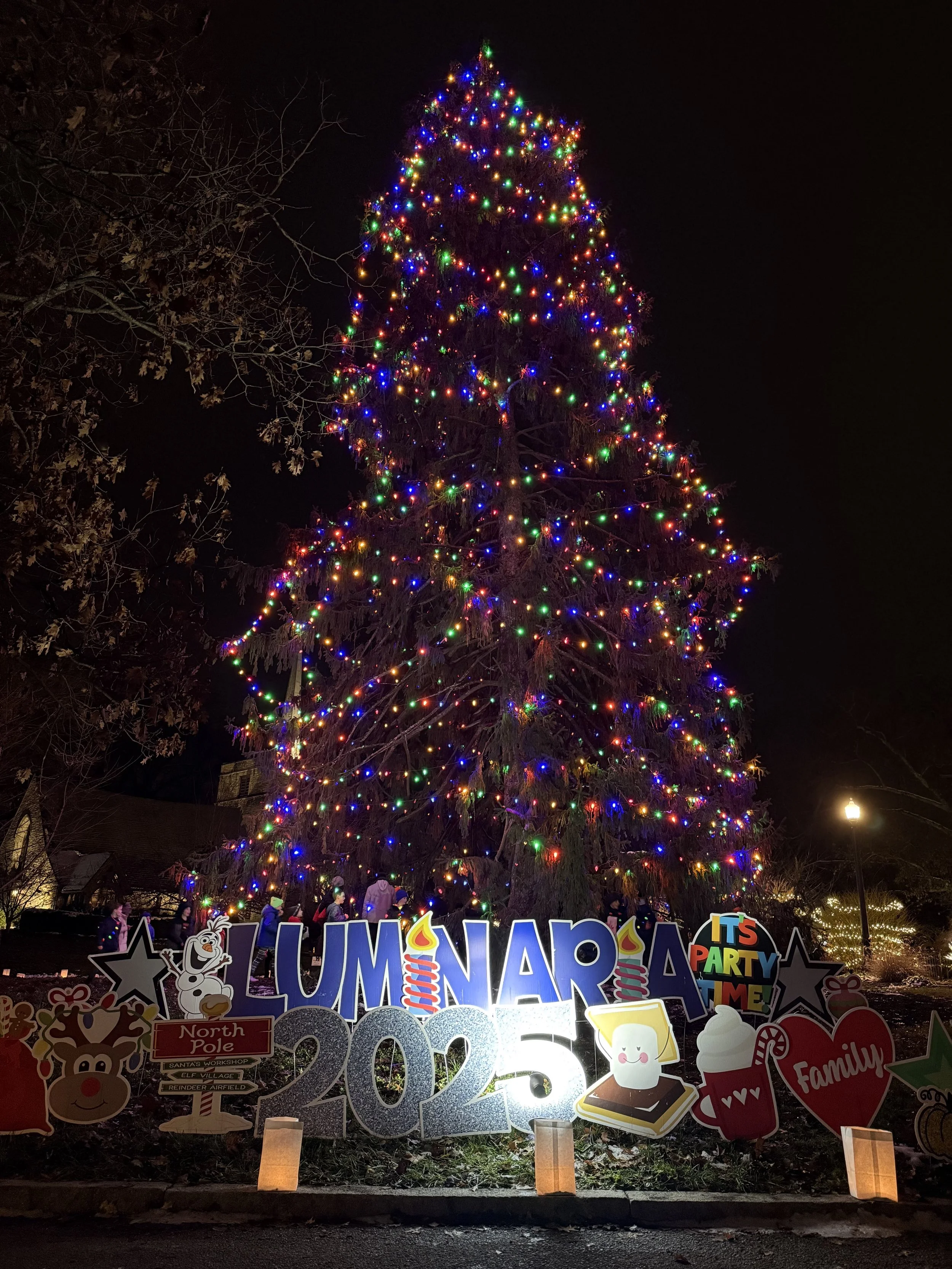 Nighttime scene of a large outdoor Christmas tree decorated with colorful lights. At the base, festive holiday signs with cartoon characters and messages, including a snowman, candy cane, and words like "LUMINARY" and "IT'S PARTY TIME!" are visible. Small lanterns illuminate the area at ground level.