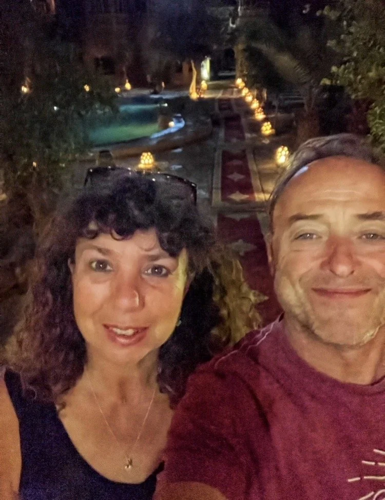 A man and woman taking a selfie outdoors at night, with a decorated pathway and outdoor pool in the background.