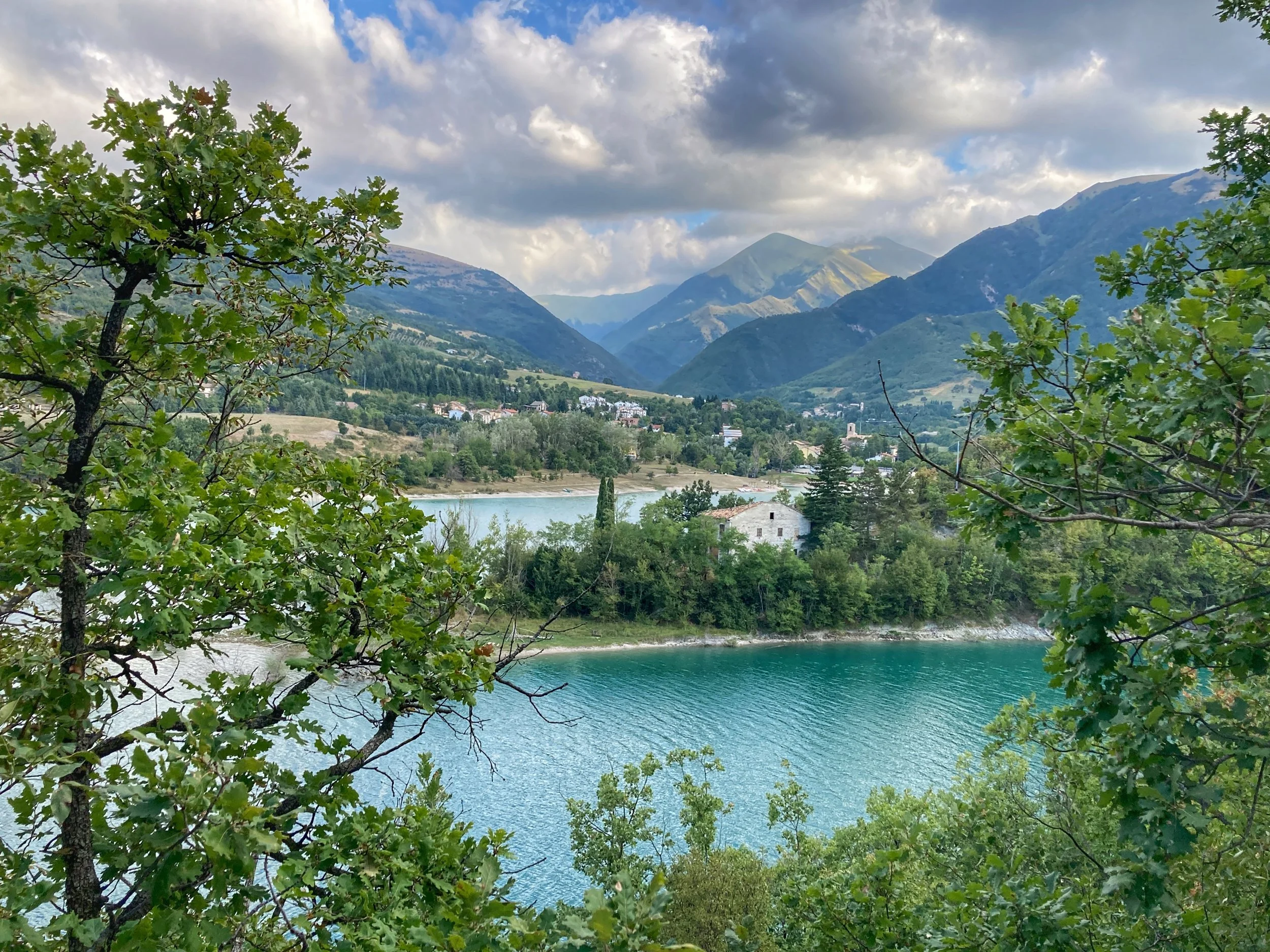 Scenic view of mountains with a village at the base, a lake in the foreground, and trees framing the scene, under a partly cloudy sky.