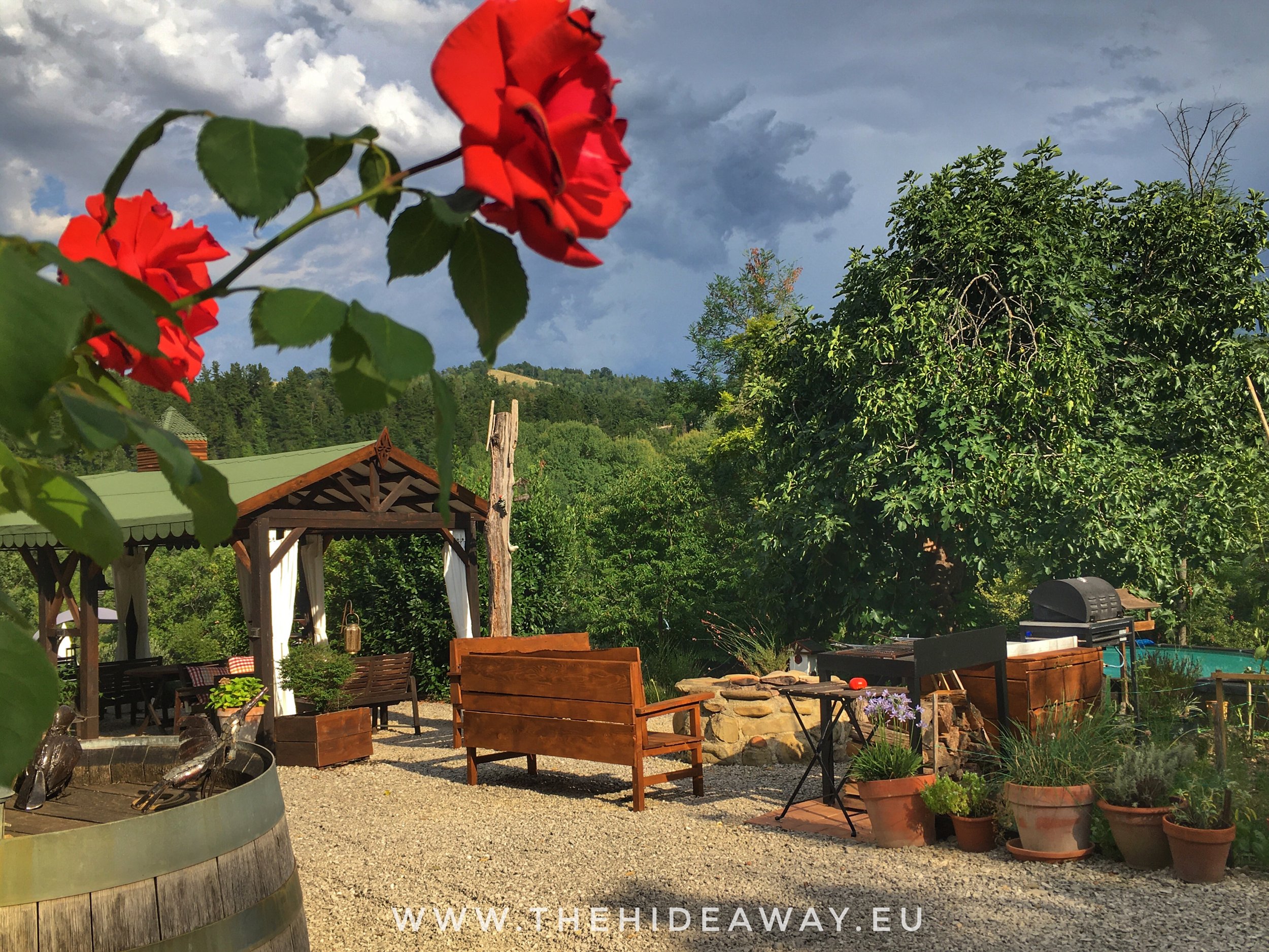 An outdoor garden patio area with wooden benches, a table, potted plants, a fire pit, and a pergola under a cloudy sky with a green hill in the background.