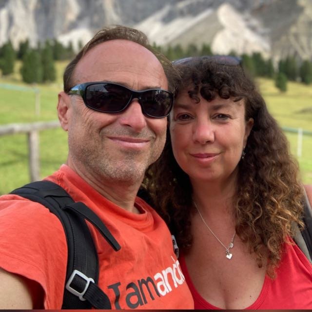 A man and woman posing outdoors in a green, mountainous landscape, both smiling at the camera.