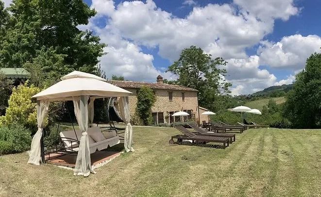 Outdoor garden with a gazebo, lounge chairs, and umbrellas on a grassy lawn, with a brick house and green trees under a partly cloudy sky.