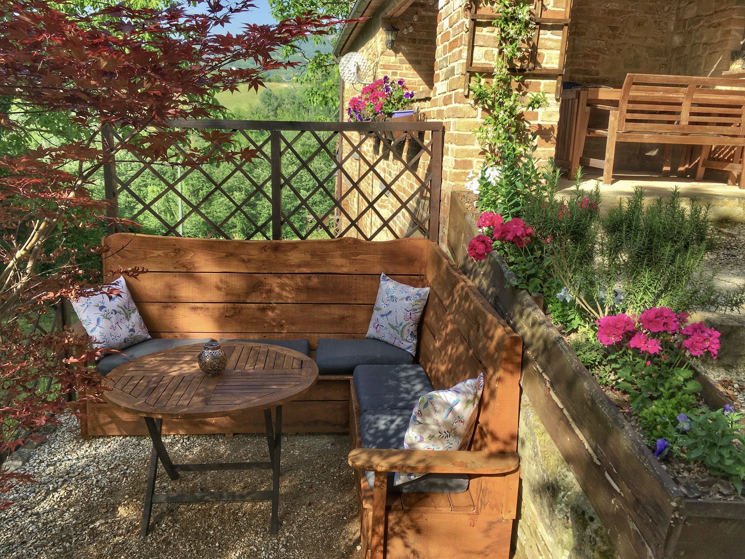 A cozy outdoor patio with wooden and metal furniture, surrounded by lush greenery and vibrant flowers on a sunny day.