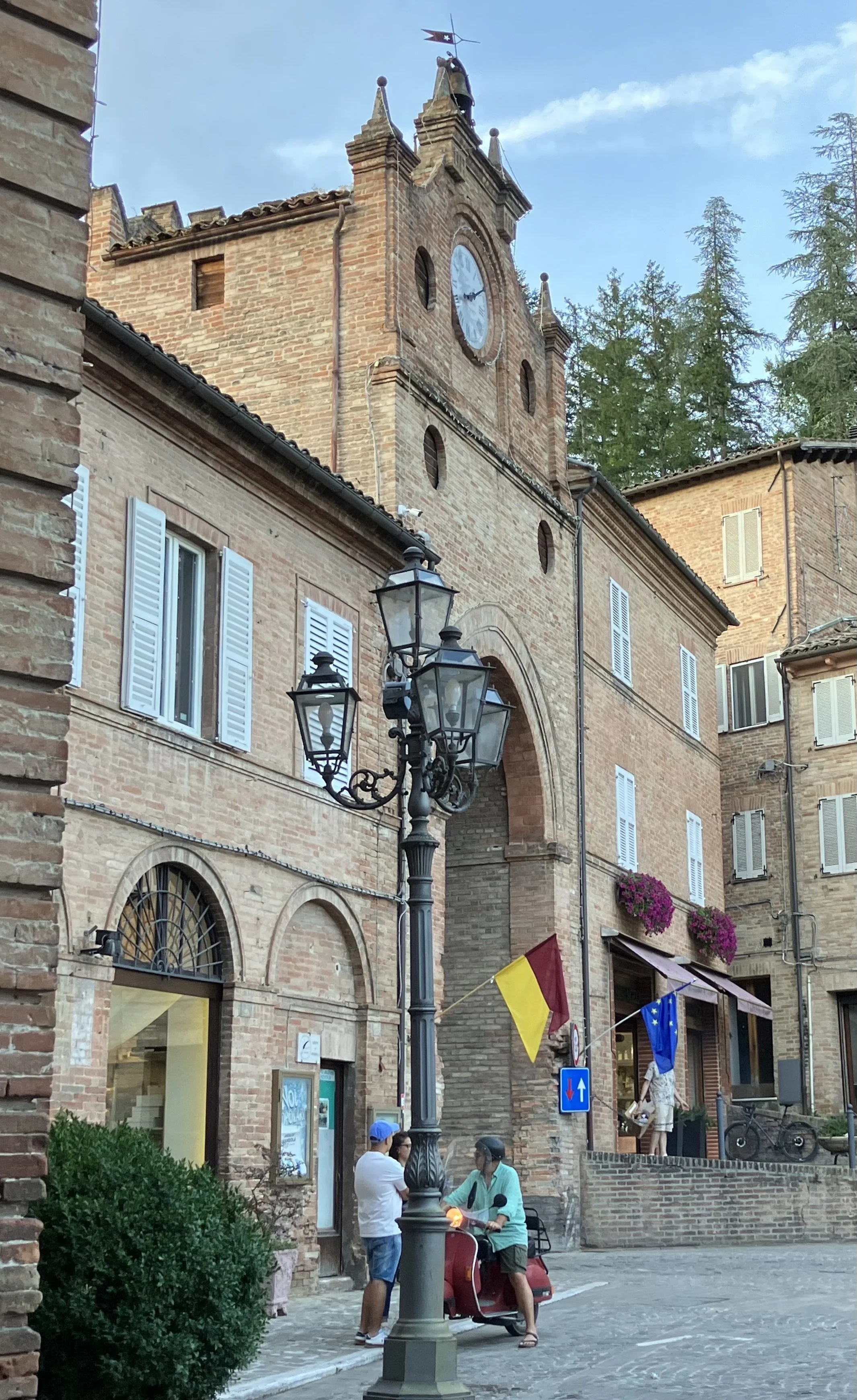 A street scene with historic brick buildings, a vintage street lamp, and flags including the Belgian and European Union flags. People are gathered around, some sitting and some standing, near a scooter on a cobblestone street.