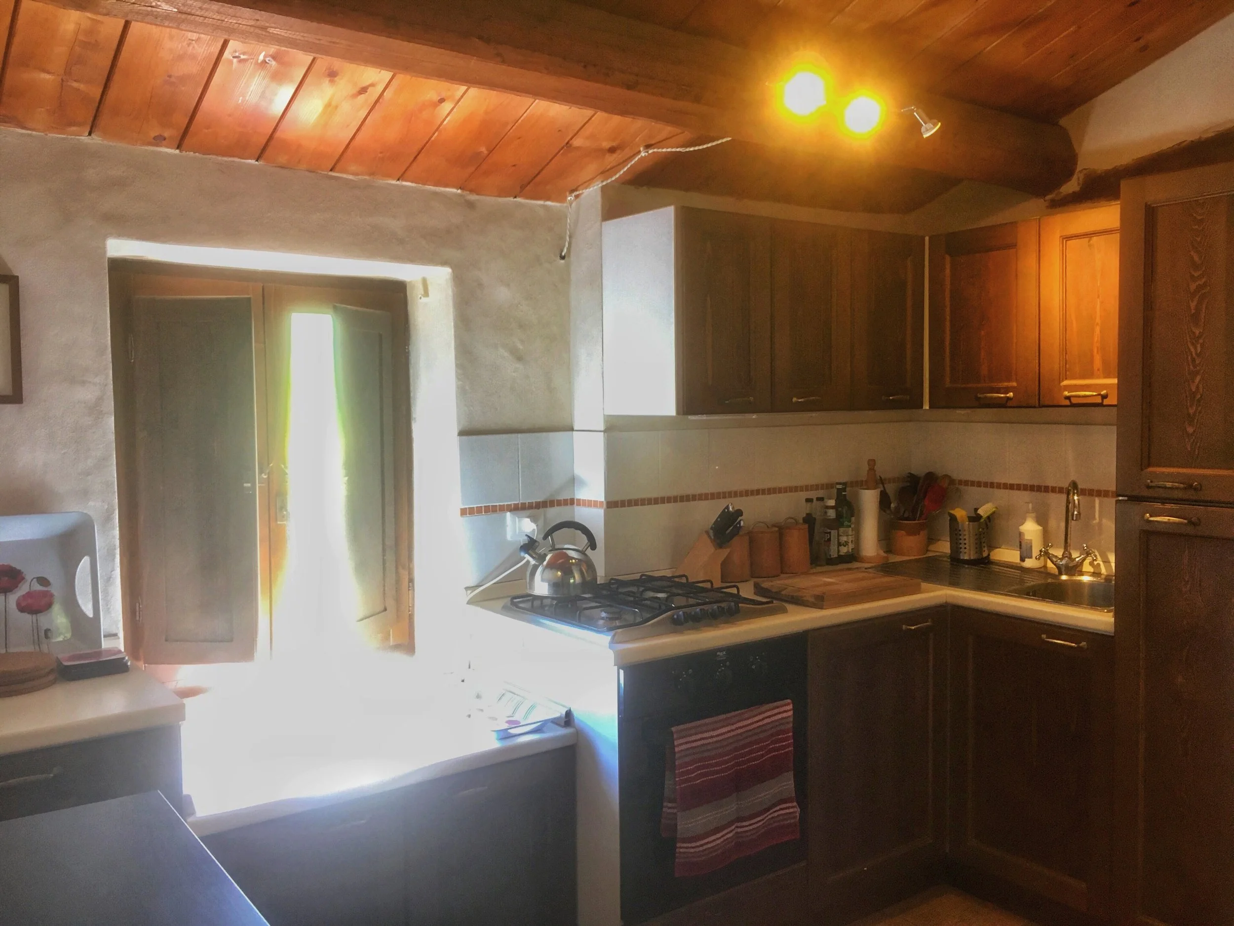 A kitchen with wooden cabinets, a countertop, and a stove, illuminated by hanging light fixtures with yellow bulbs. There is a window above the sink, and various kitchen items on the counter.